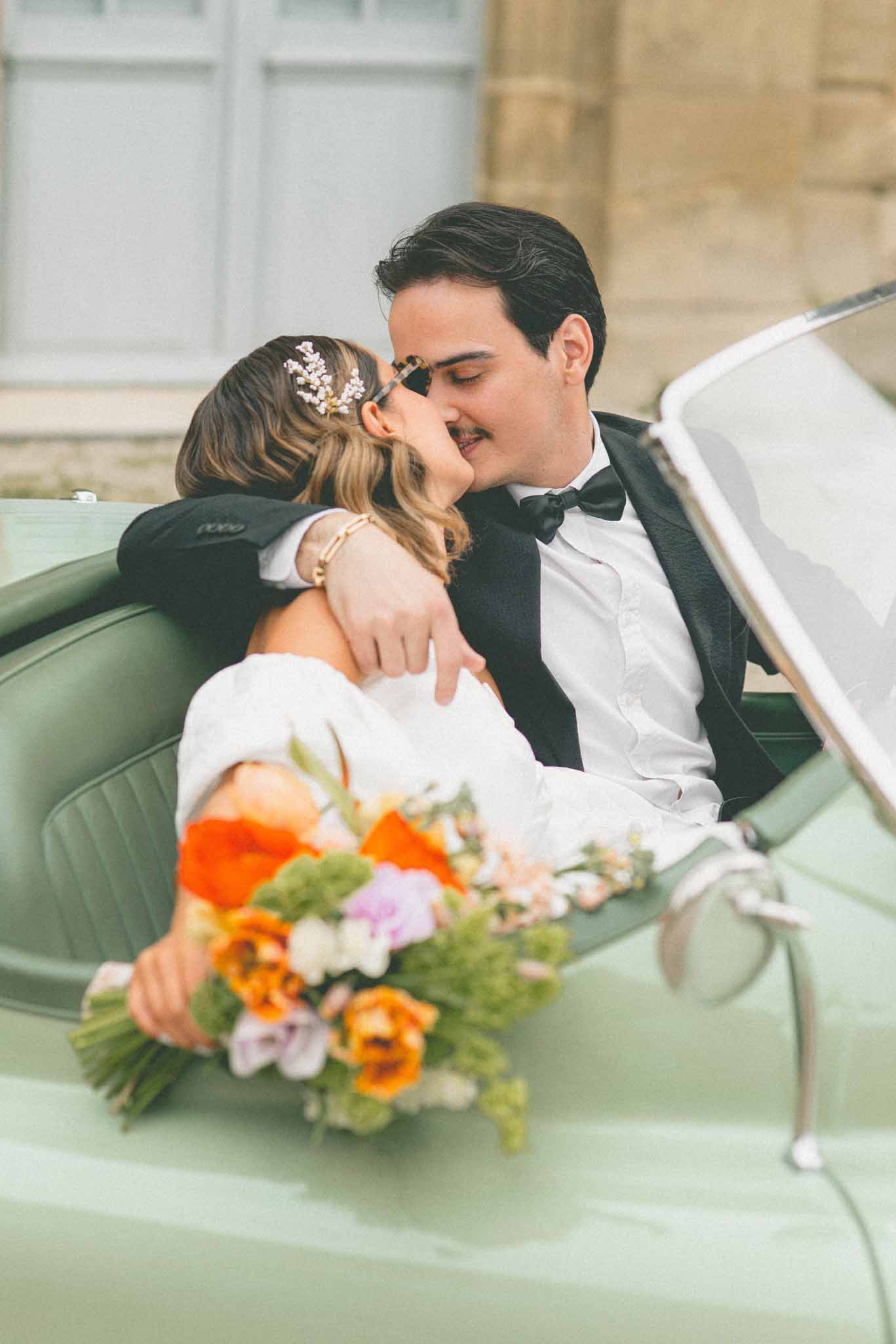 Bride and groom kissing in mint green vintage convertible, bride holding orange poppy and lavender bouquet