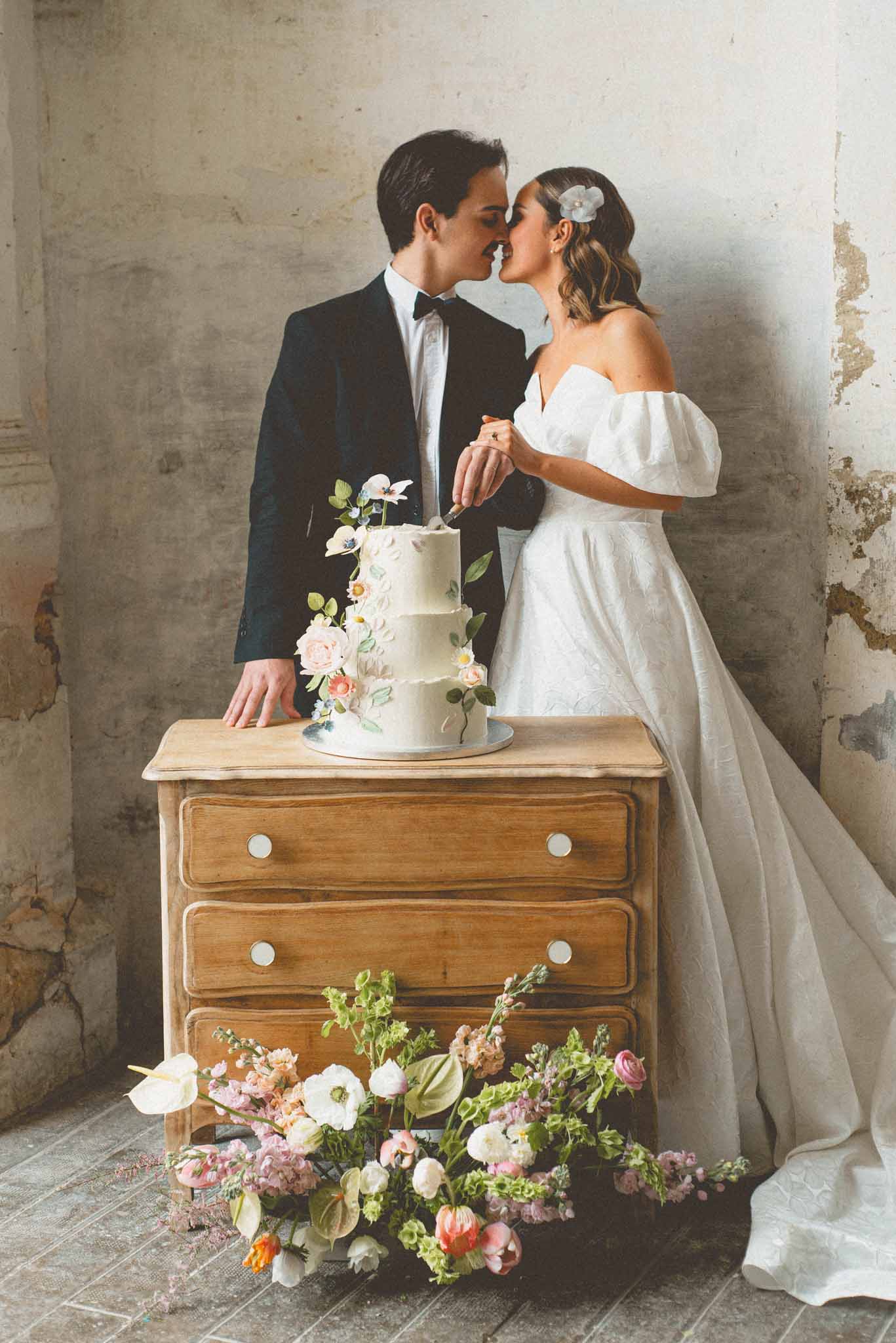 A couple cuts their wedding cake in what appears to be an indoor space with exposed, aged plaster walls, suggesting a historic or rustic venue interior. The groom wears a navy tuxedo with a black bow tie, and the bride wears an off-the-shoulder white ball gown with voluminous puff sleeves and a full skirt, accented by a floral hair clip. The three-tier white wedding cake sits on a natural wood chest of drawers and is decorated with sugar flowers in blush pink, coral, and white tones with green foliage details. A loose floral arrangement of blush ranunculus, white anemones, coral tulips, anthurium leaves, and pink stock flowers is placed directly on the stone tile floor in front of the dresser. The couple leans in toward each other as they hold the cake knife together, shot as a mid-range portrait with full-length framing.