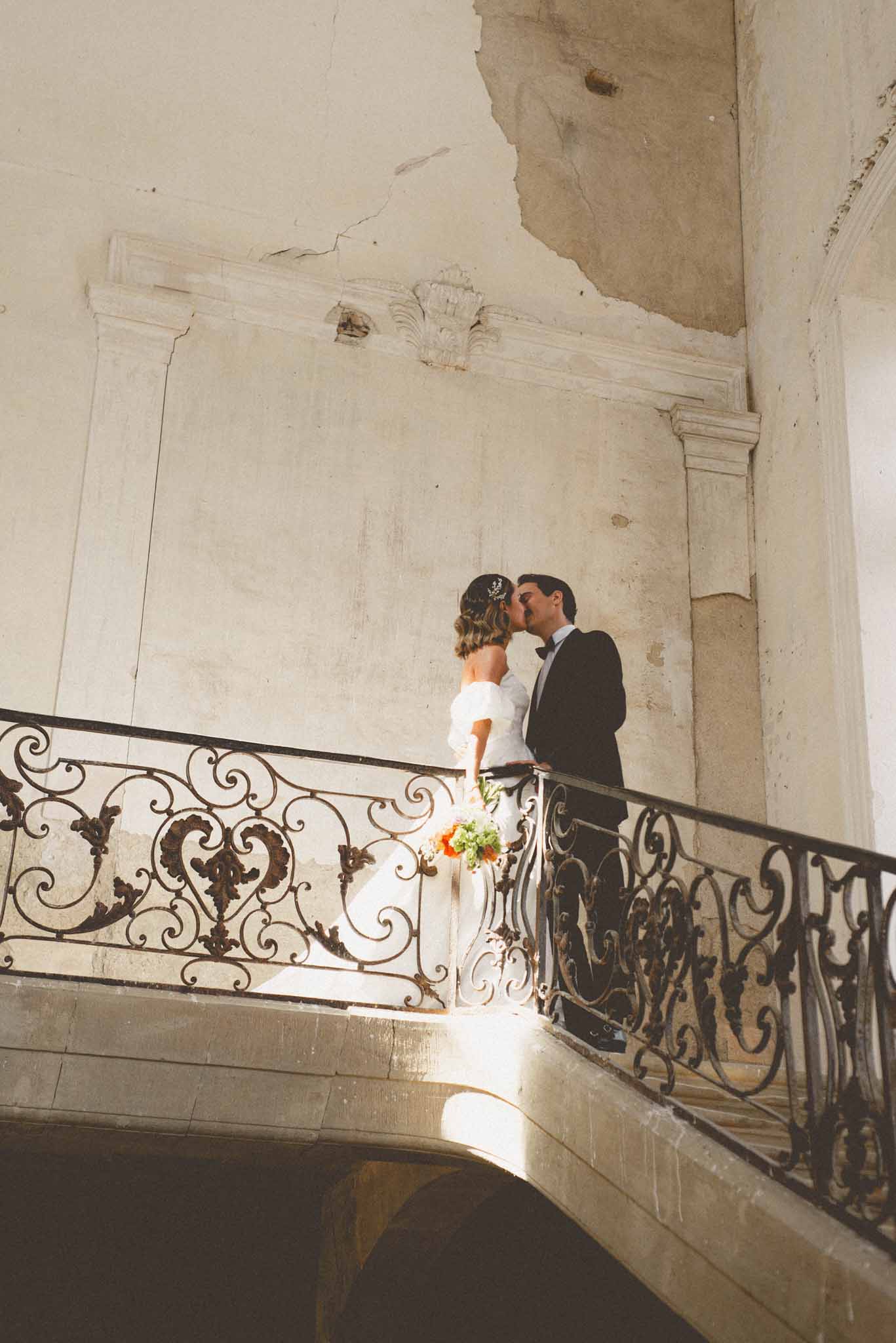 A couple portrait taken indoors on the landing of a curved stone staircase inside what appears to be a French château or historic manor, featuring ornate wrought-iron scrollwork balustrades. The bride and groom are kissing; the bride wears an off-the-shoulder white ball gown with voluminous ruffled sleeves and a floral hair accessory, and holds a bouquet with orange and coral blooms and green foliage. The groom is dressed in a black tuxedo with a bow tie. Natural light streams in from below, casting a warm patch of light on the stone landing. The interior walls show aged, peeling plasterwork with classical molding details, contributing to a worn, historic aesthetic. The composition is a wide portrait shot taken from a low angle, looking up toward the couple.