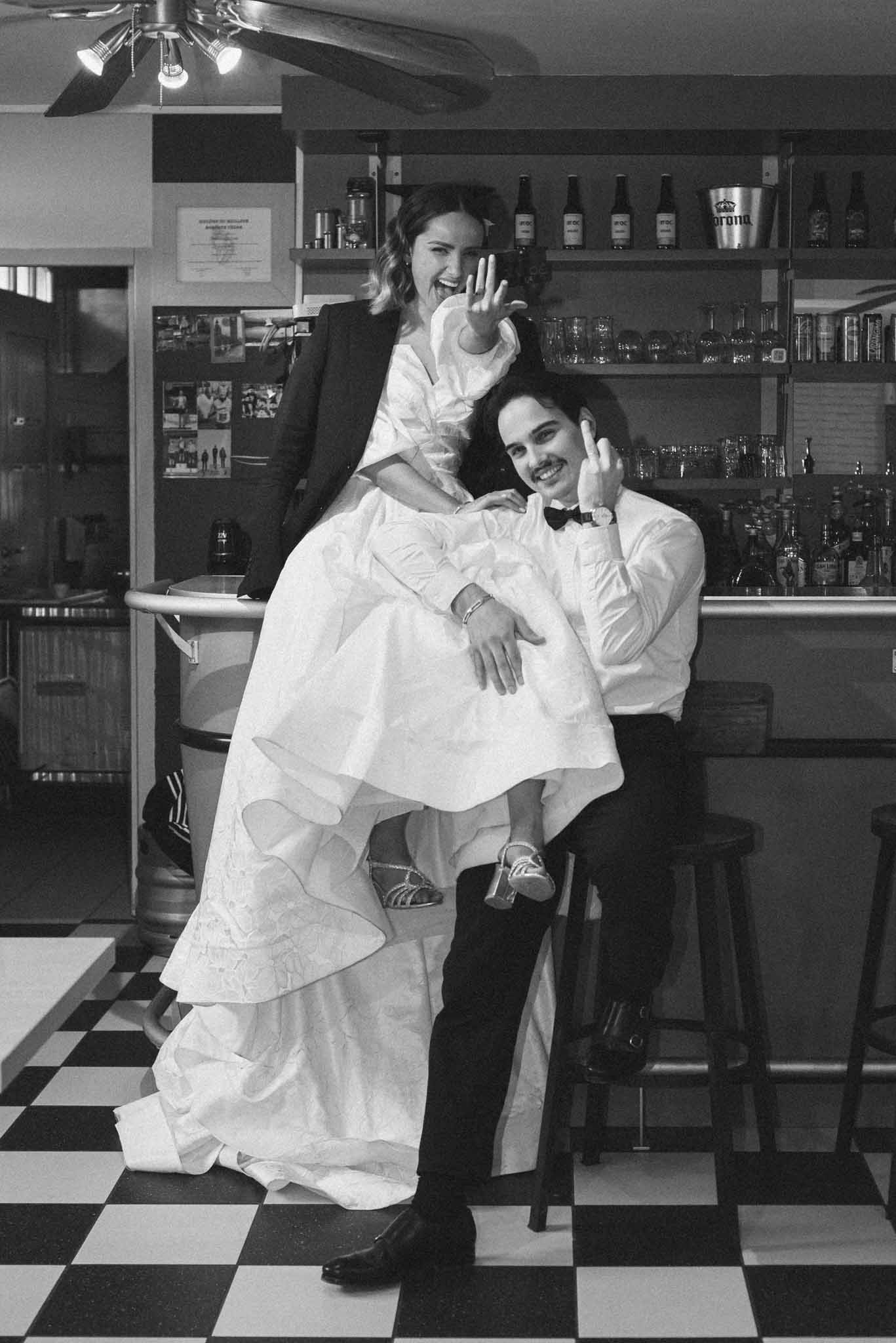 Black and white portrait of bride sitting on groom's lap at a bar, both laughing and showing wedding ring