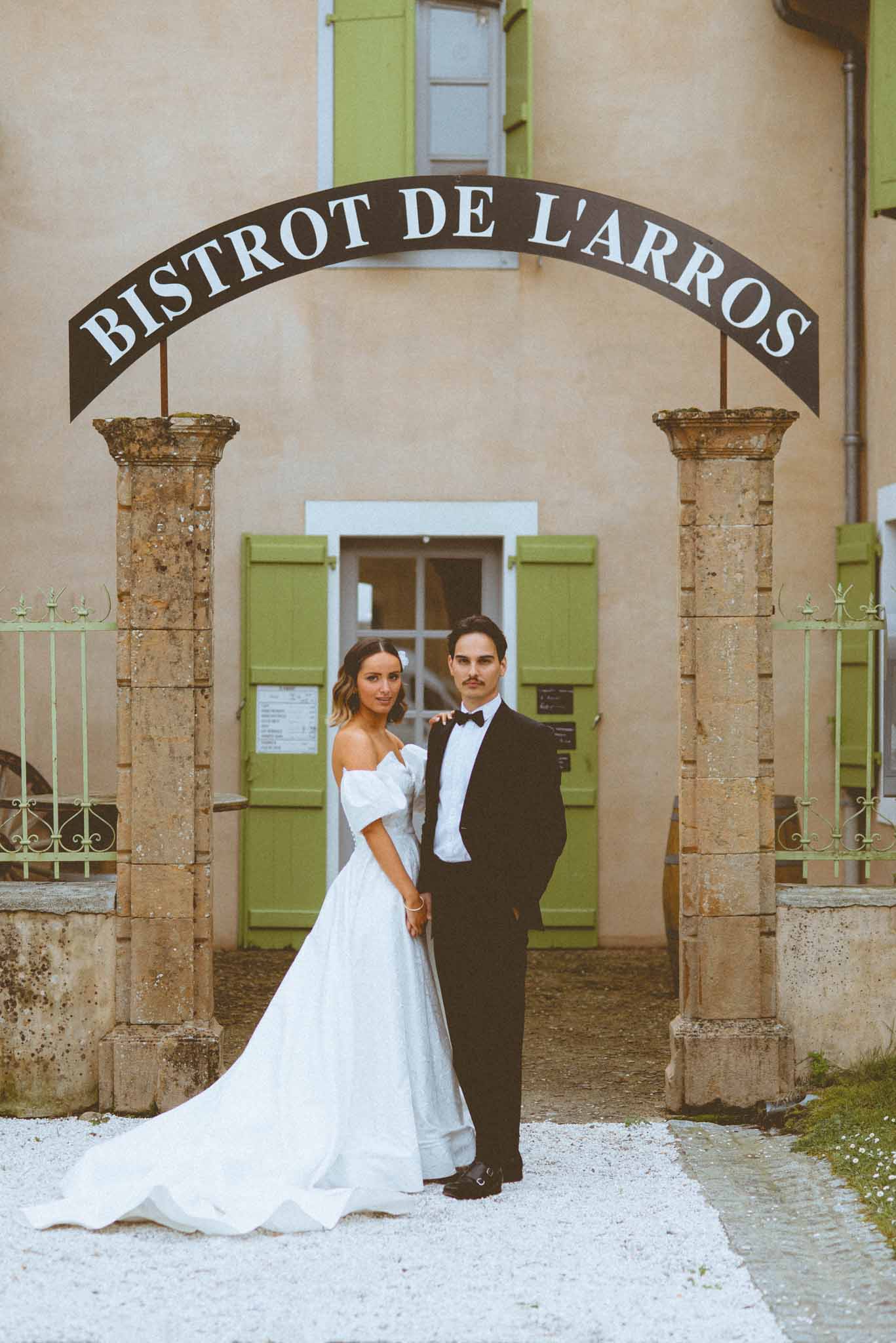 Couple stands between stone pillars under Bistrot de l'Arros iron sign with sage gates and ochre facade