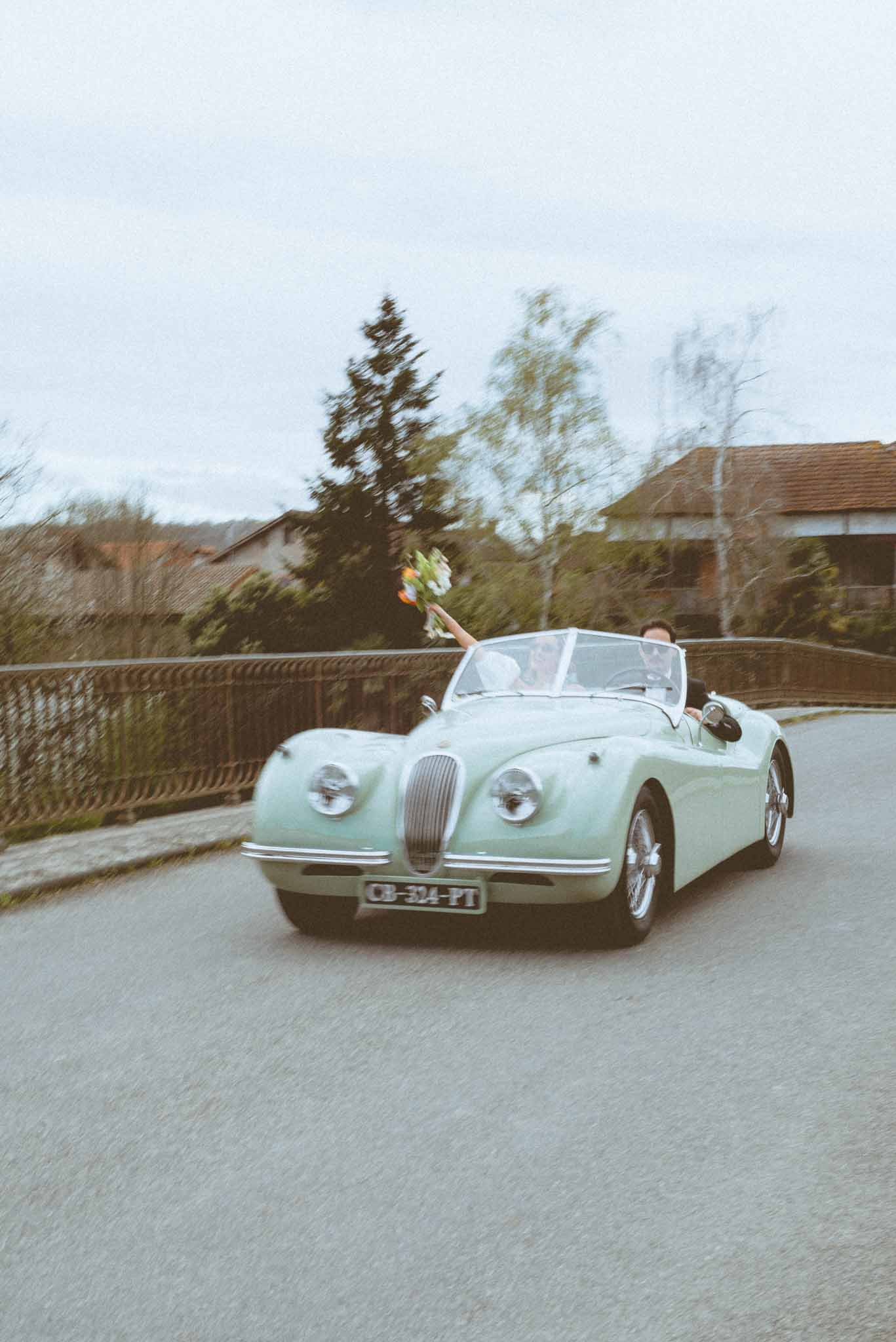 A couple is driving along a rural road in a mint green vintage convertible sports car, consistent with a classic Jaguar XK120, with the top down. The bride, dressed in white, is raising her bouquet of mixed blooms with orange and green tones above the windscreen in a celebratory gesture, while the groom drives beside her. The shot is taken from a low front-facing angle as the car approaches, giving a dynamic, motion-filled quality with slight background blur suggesting movement. The setting is an outdoor country road flanked by an iron railing and residential buildings in the background, consistent with a small French village.