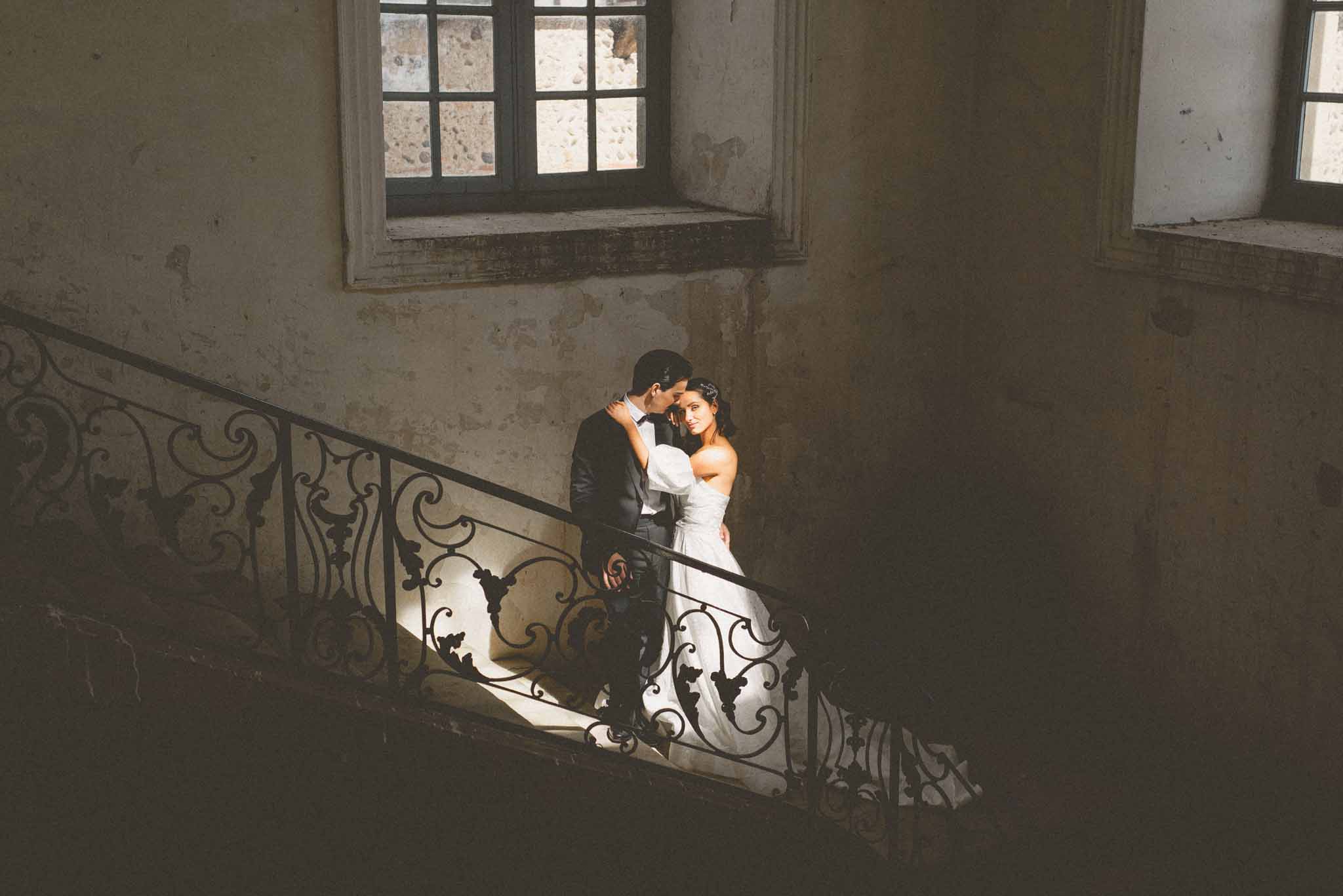 Bride in strapless white ballgown and groom in dark suit posing on ornate wrought-iron staircase in French chateau
