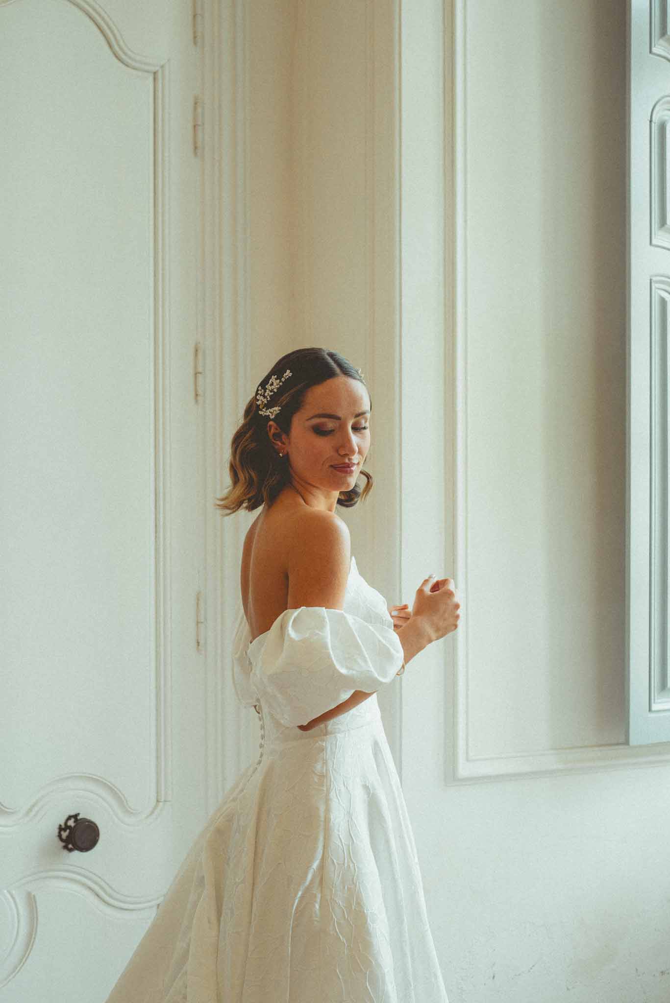 Bride in off-the-shoulder puff sleeve brocade gown adjusting bracelet in front of ornate white panelled doors