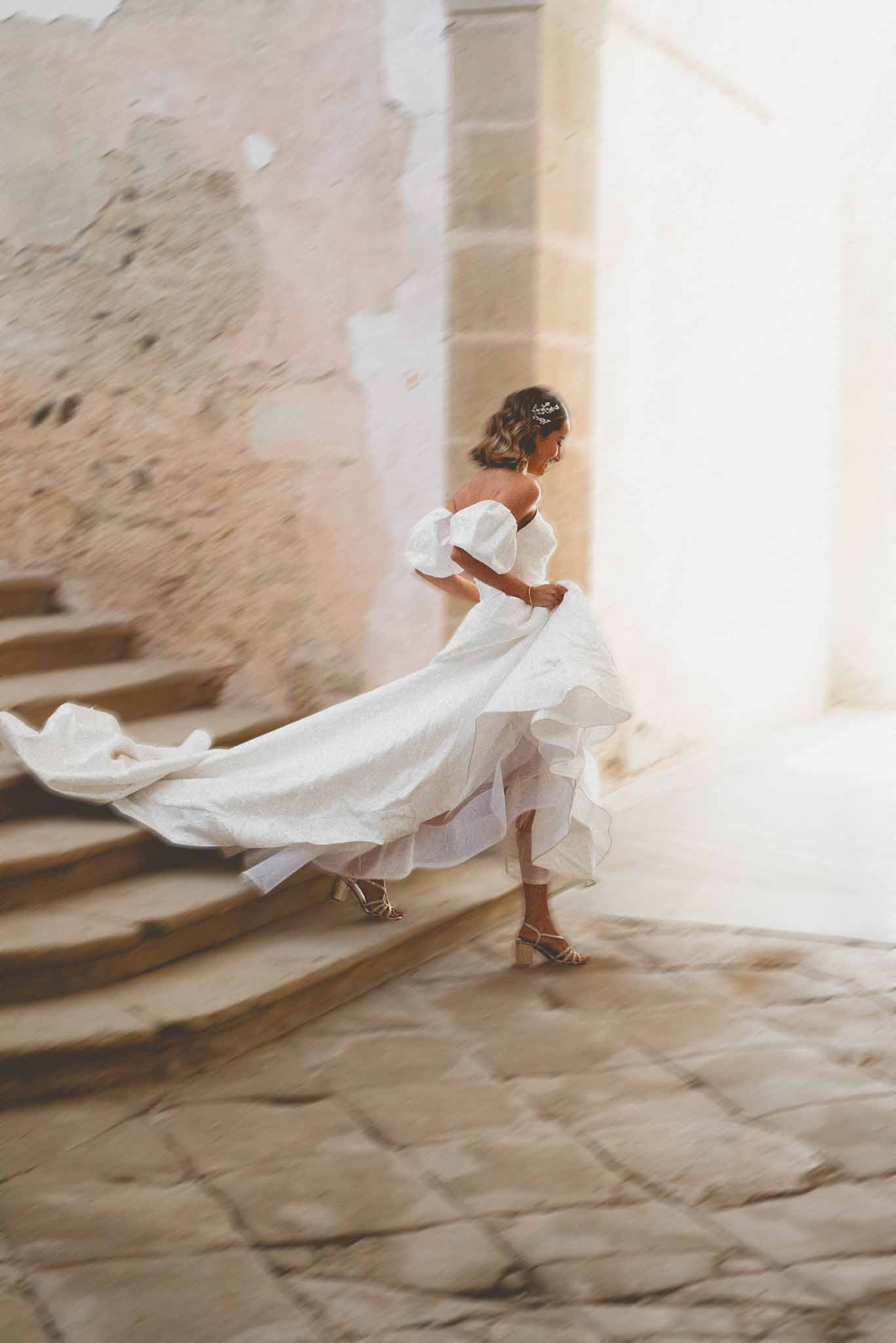 Bride mid-turn in off-shoulder puff-sleeve gown with layered ruffled train and gold sandals on cobblestone