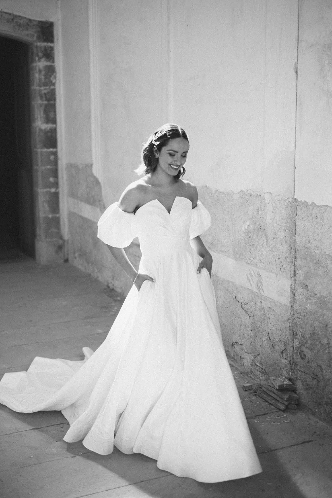 Black and white portrait of bride in ball gown with puff sleeves and hands in pockets against aged plaster wall