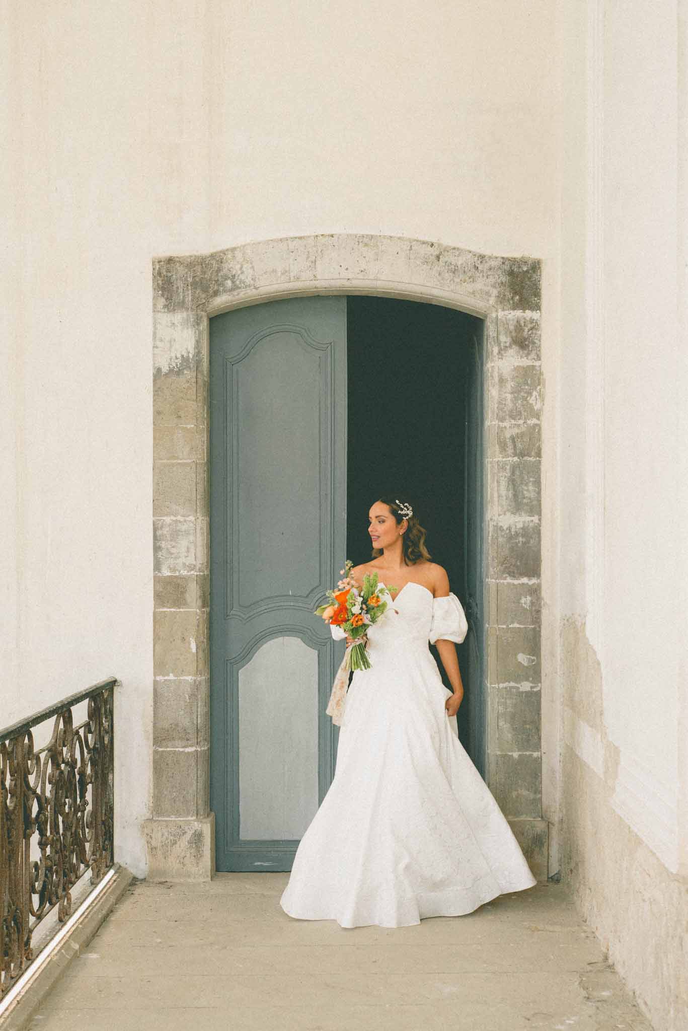 Bride in puff-sleeve ballgown with orange ranunculus bouquet before slate-blue panelled chateau doorway