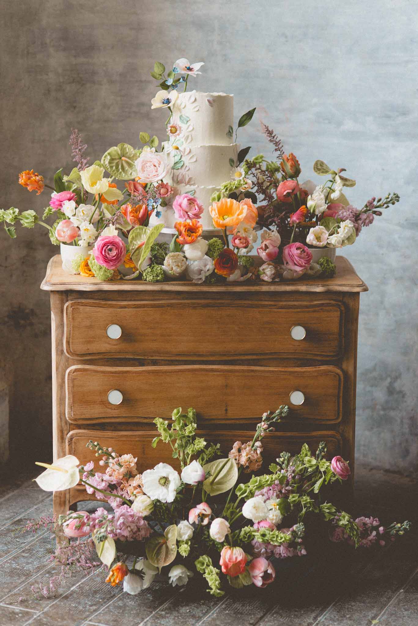 A styled detail shot featuring a two-tier white wedding cake with textured buttercream frosting and delicate sugar flower appliqués, displayed on top of a vintage honey-toned wooden chest of drawers with round white knobs. The cake is surrounded by a dense, garden-style floral arrangement in a vibrant palette of hot pink ranunculus, orange poppies, coral tulips, blush roses, white anemones, yellow blooms, anthurium leaves, and greenery. An additional loose floral arrangement spills across the tiled floor in front of the dresser, incorporating white anemones, pink ranunculus, lavender stock, peach tulips, and mixed foliage. The overall styling theme is bohemian and maximalist, with a rich mix of colors and textures against a muted grey concrete-style wall. The composition is a straight-on medium shot capturing the full dresser from floor to top.