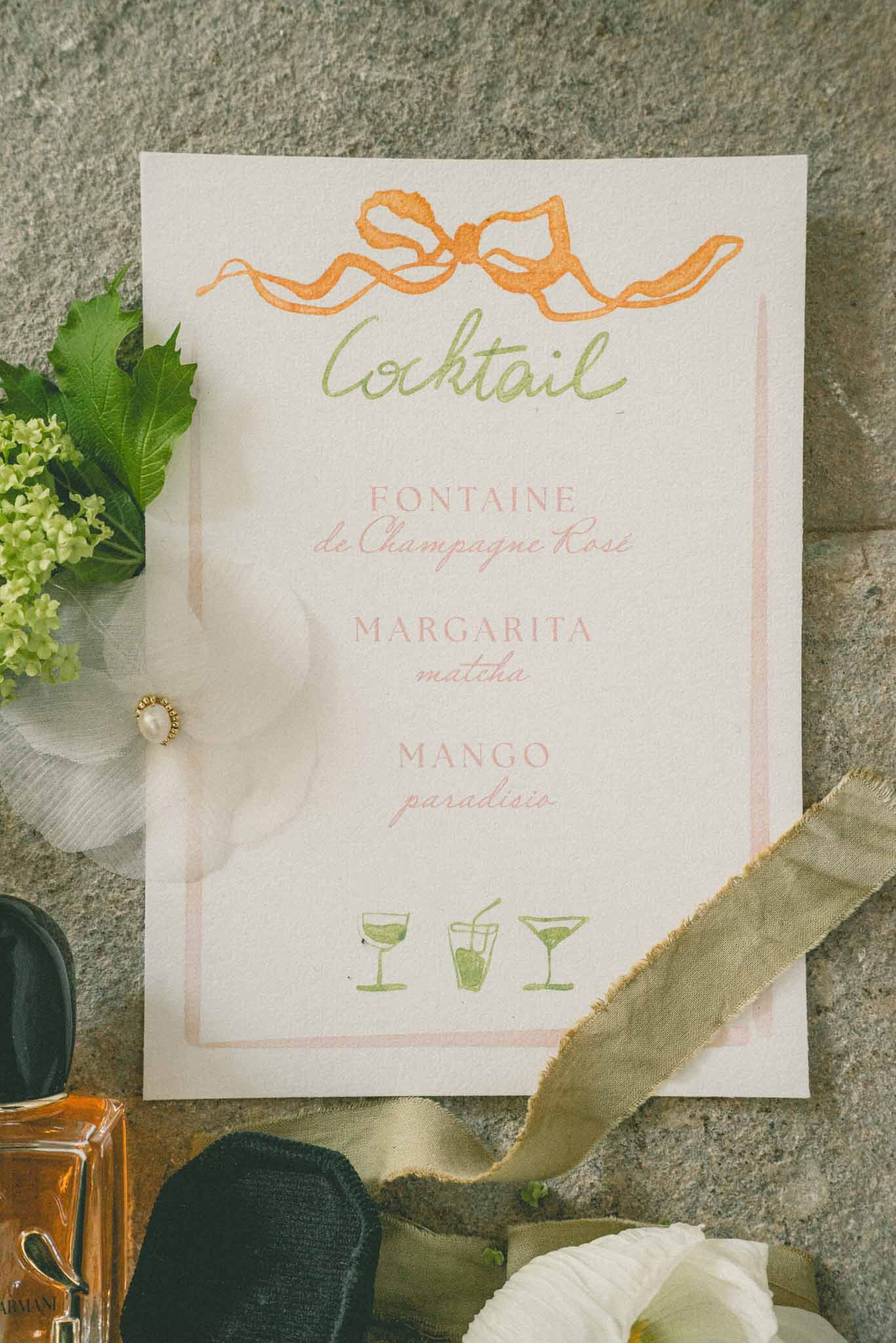 Flat lay of illustrated cocktail menu card with bridal accessories, perfume, and green ribbon on stone surface