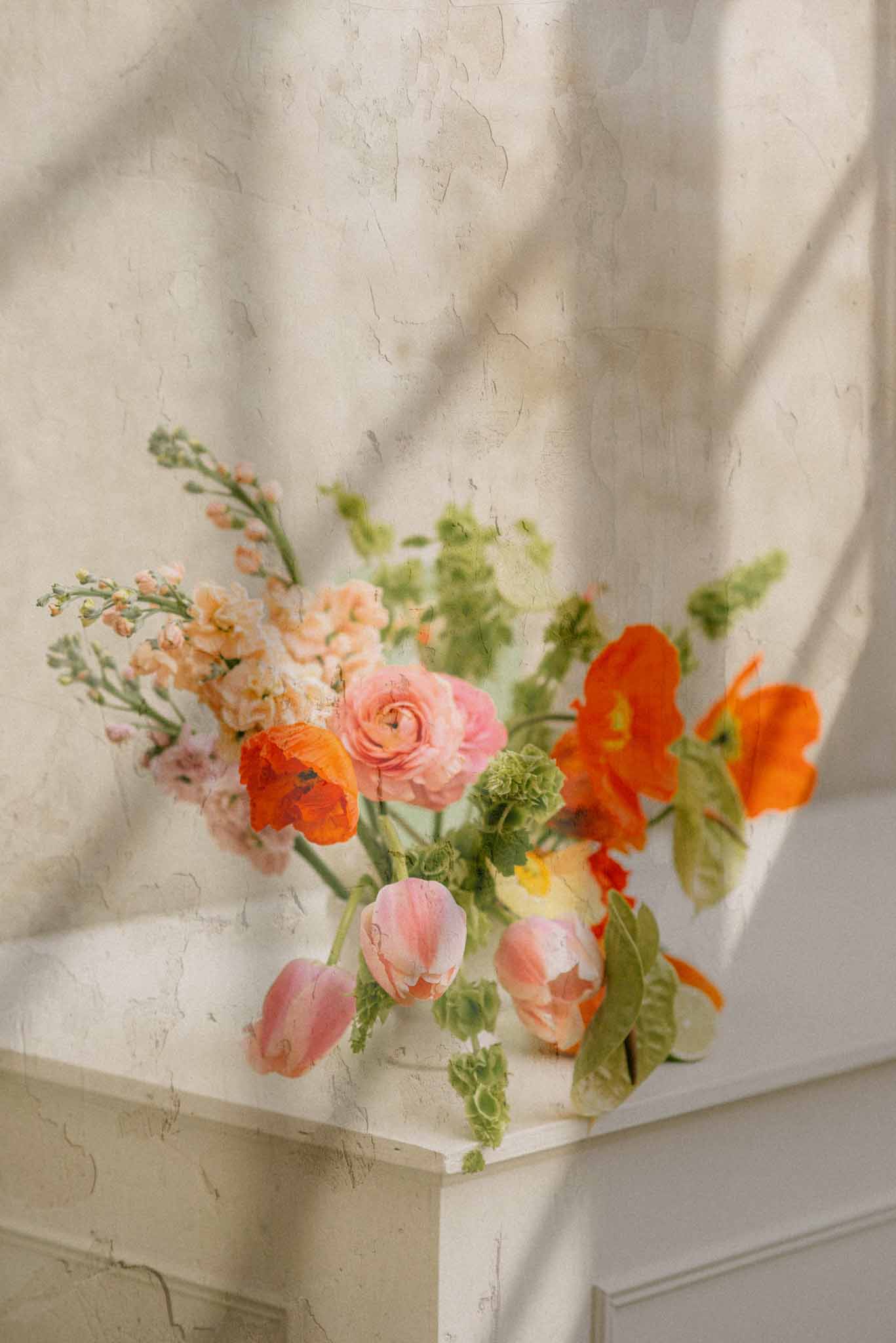 Loose arrangement of orange poppies, blush ranunculus, and pink tulips on stone mantelpiece in dappled light
