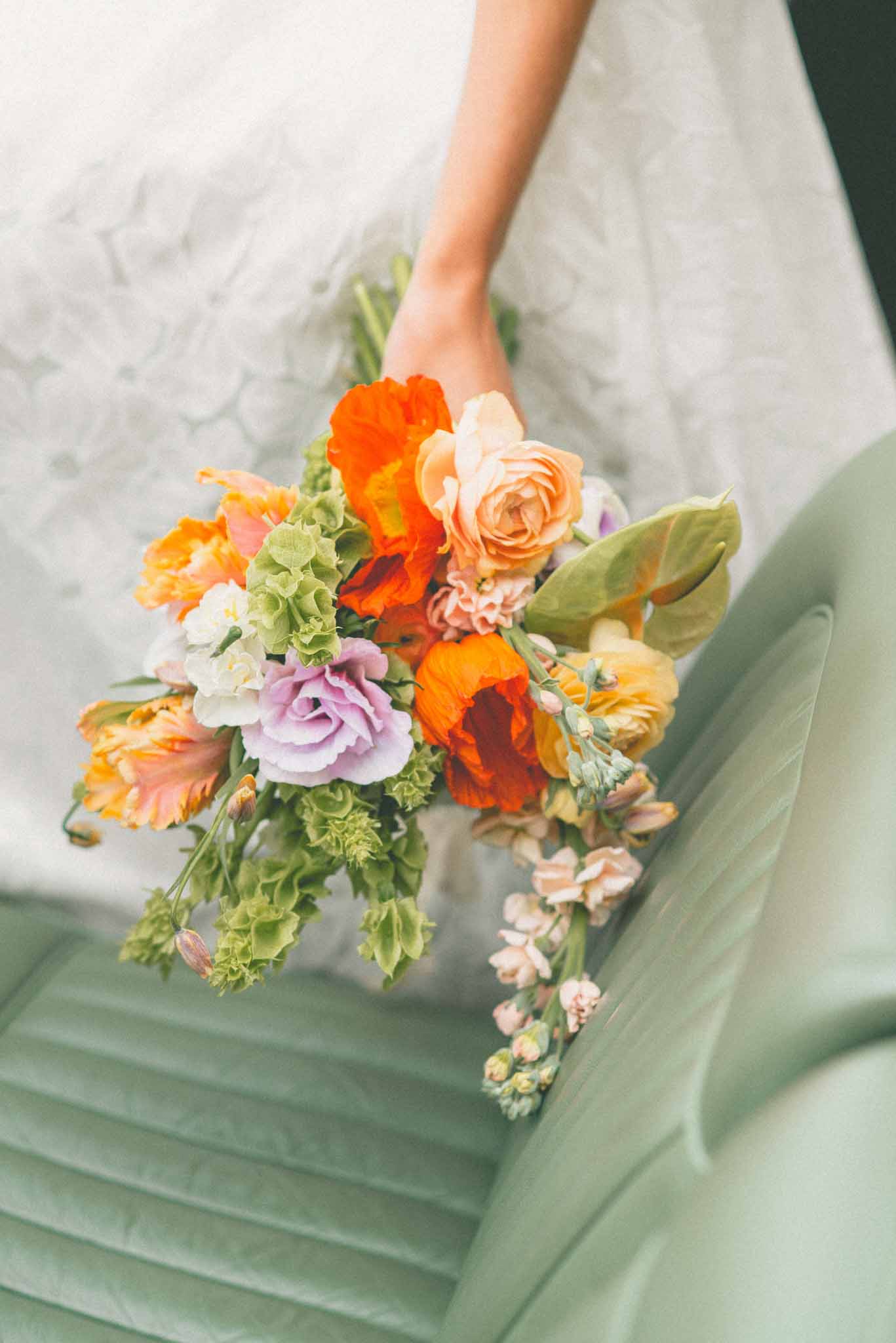 A close-up detail shot of a bridal bouquet held by a bride wearing a white lace dress. The bouquet is a loose, garden-style arrangement featuring orange Iceland poppies, peach garden roses, lavender lisianthus, yellow ranunculus, white sweet peas, green bell-shaped blooms, blush stock flowers, and trailing green foliage with leaves. The composition is shot from above at a slight angle, with the bouquet resting against what appears to be a sage green velvet chair or sofa, creating a soft color contrast with the vibrant warm tones of the florals. The overall palette is bold and colorful, mixing warm oranges and peaches with cooler lavender and sage green accents in a relaxed, unstructured style.