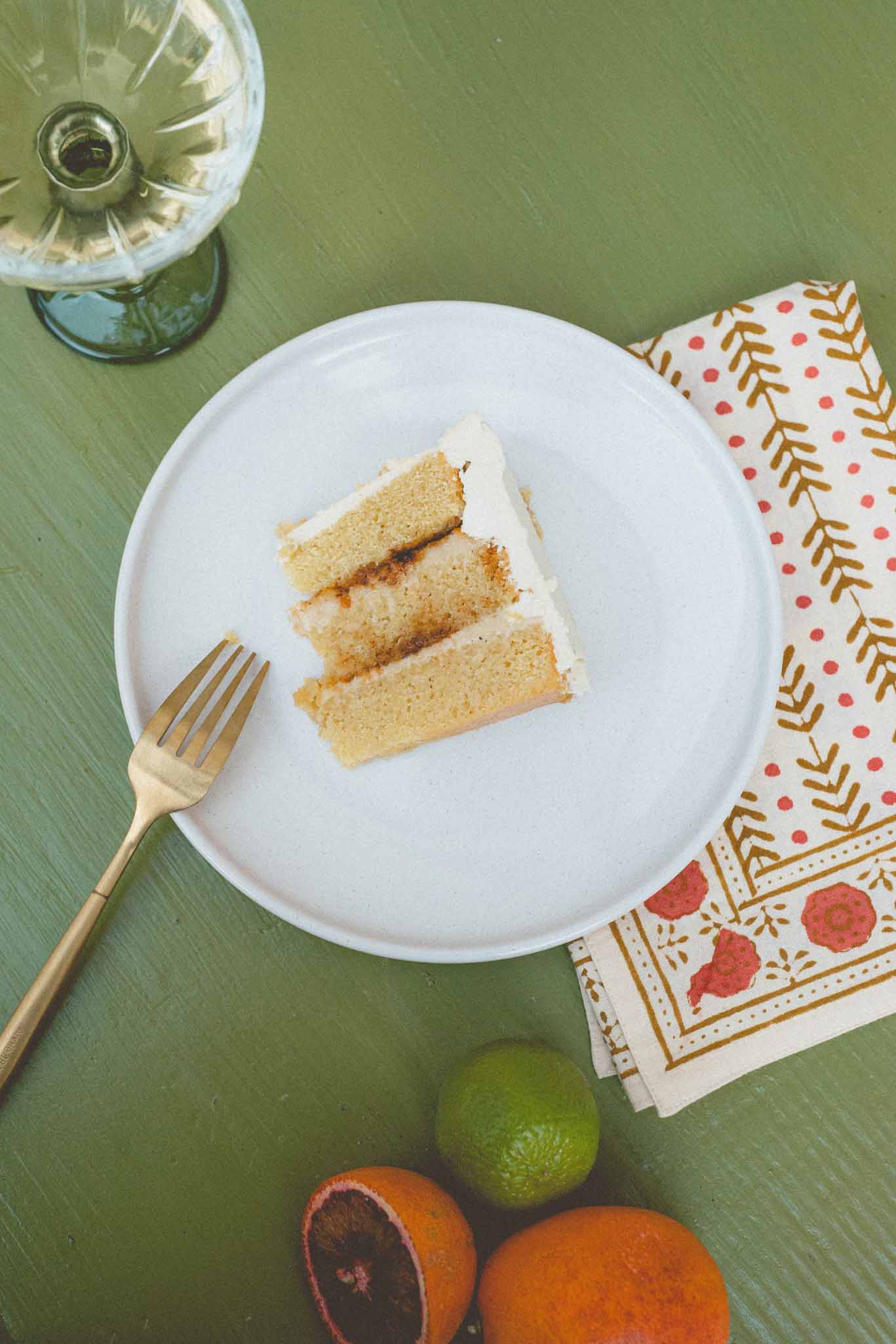 Overhead detail of cake slice on white plate with gold fork, citrus fruits, and botanical napkin on sage green surface