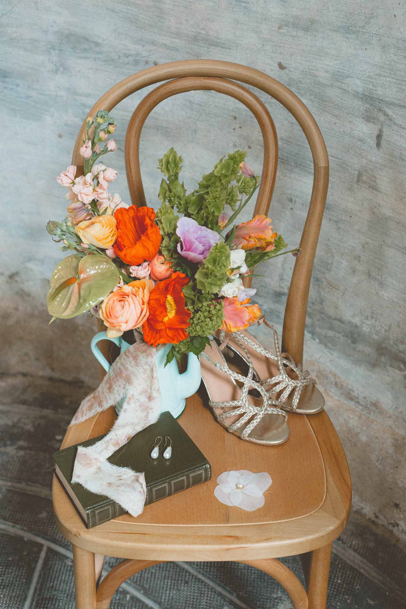 Bridal flat lay on bentwood chair with orange poppy and peach rose bouquet, gold strappy sandals, and pearl drop earrings
