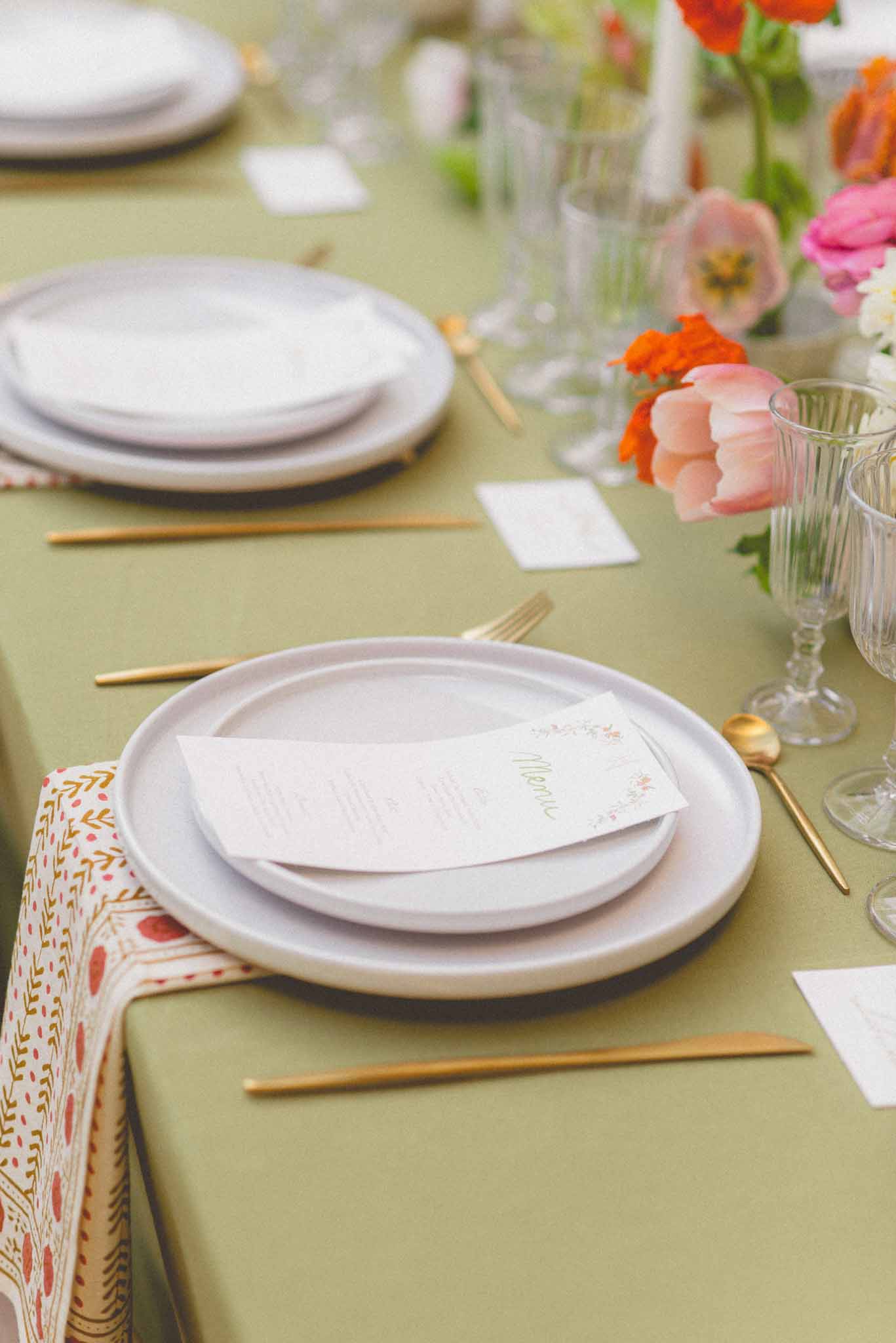 Close-up detail shot of a wedding reception table setting featuring a sage green linen tablecloth and stacked matte grey ceramic plates with a printed menu card on top. Gold flatware is laid flat on the tablecloth rather than upright, and ribbed clear glassware is placed to the right of each setting. A block-print napkin in cream with red and gold botanical motifs is folded to the left of the plates. Small white place cards are visible between settings. The floral centerpiece running along the table includes blush pink tulips, orange poppies or ranunculus, and hot pink blooms, creating a vivid, colorful contrast against the muted sage green linen. The overall styling palette combines sage green, gold, and warm multicolored florals for a fresh, garden-inspired reception aesthetic.