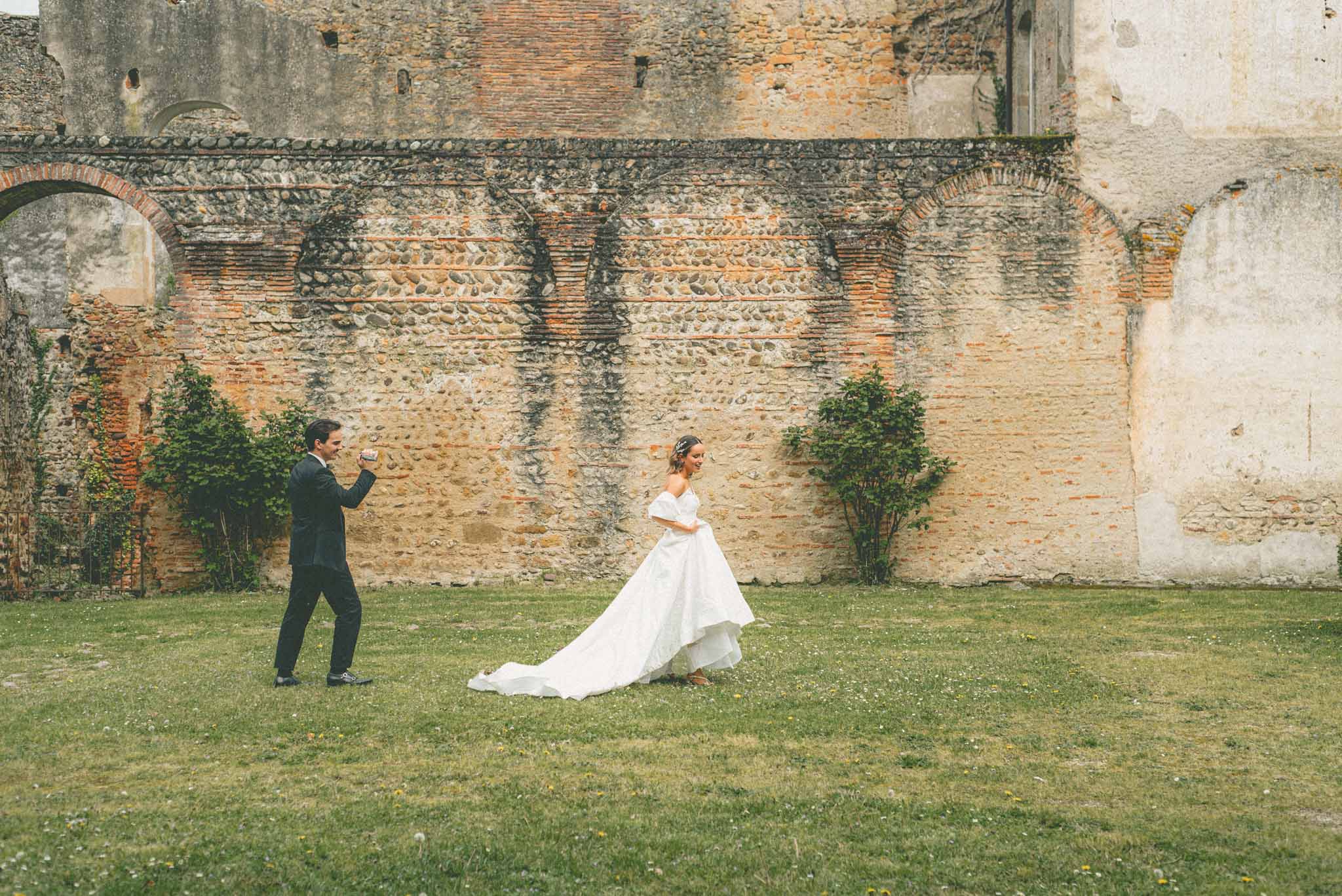 A couple portrait shot outdoors on a lawn in front of ancient stone and brick ruins with arched architectural details. The bride wears an off-the-shoulder white ball gown with a long cathedral train, and she is laughing and walking forward while lifting the front of her skirt. The groom, dressed in a dark navy suit, stands behind her in a playful celebratory pose with his fist raised. The setting has a rustic, historic character with the weathered stone wall featuring decorative brick arching as a backdrop. The image is a wide shot capturing the full length of both figures and the expanse of the lawn.