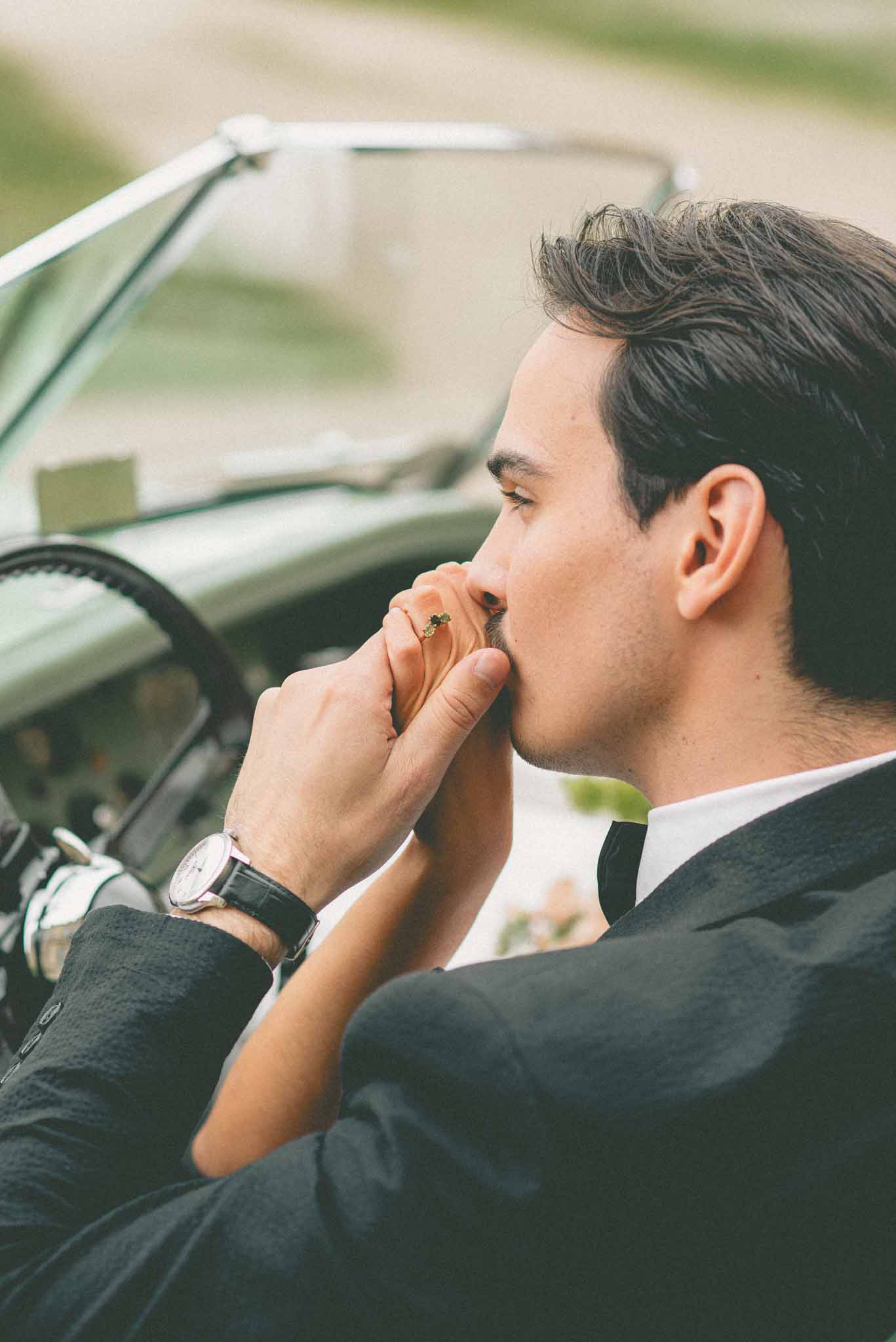 Groom in navy suit kissing partners hand inside vintage mint-green convertible with emerald ring visible