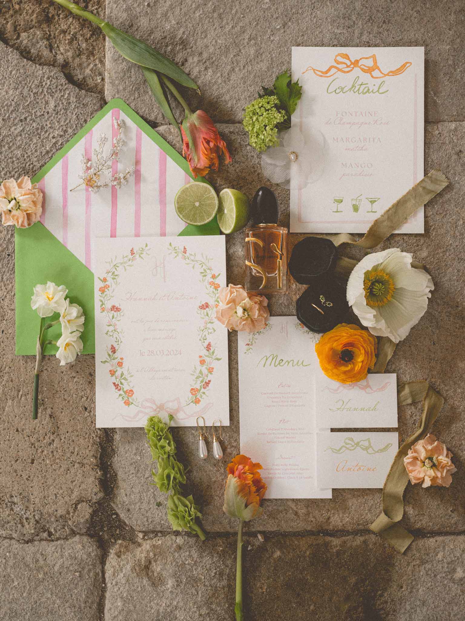 Flat lay of colorful wedding stationery suite with orange ranunculus, green envelope, cocktail menu, and ring box on stone...