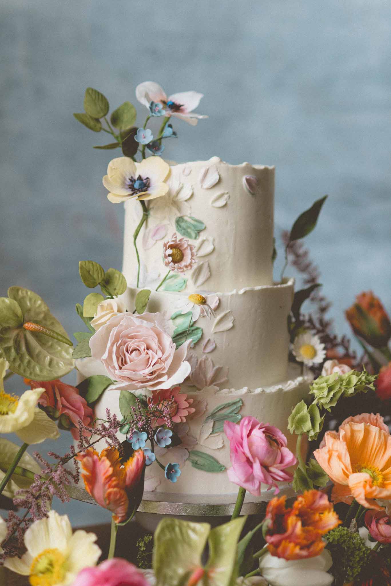 Three-tier hand-painted botanical cake with sugar flowers surrounded by parrot tulips magenta ranunculus and garden roses