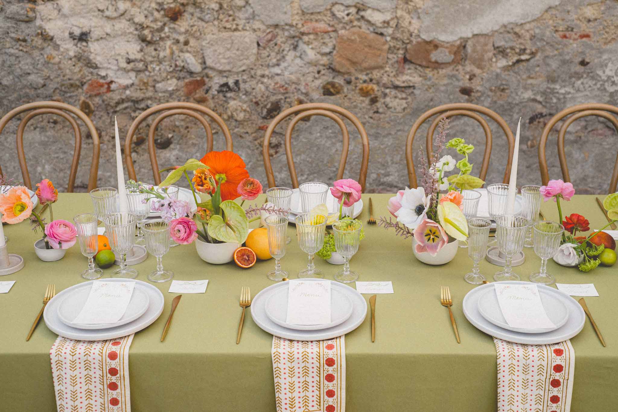 Colorful wedding tablescape with orange poppies, pink ranunculus, citrus fruits, and gold flatware on olive linen