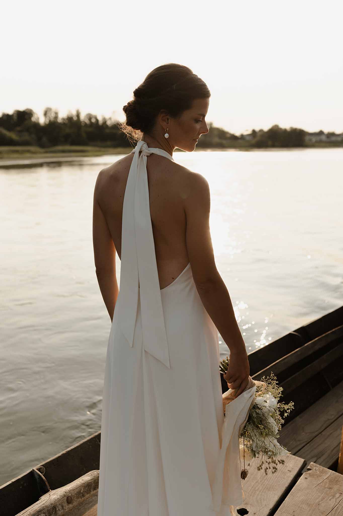 Bride in backless halter dress holding a dried grass bouquet on a wooden dock overlooking a river at golden hour
