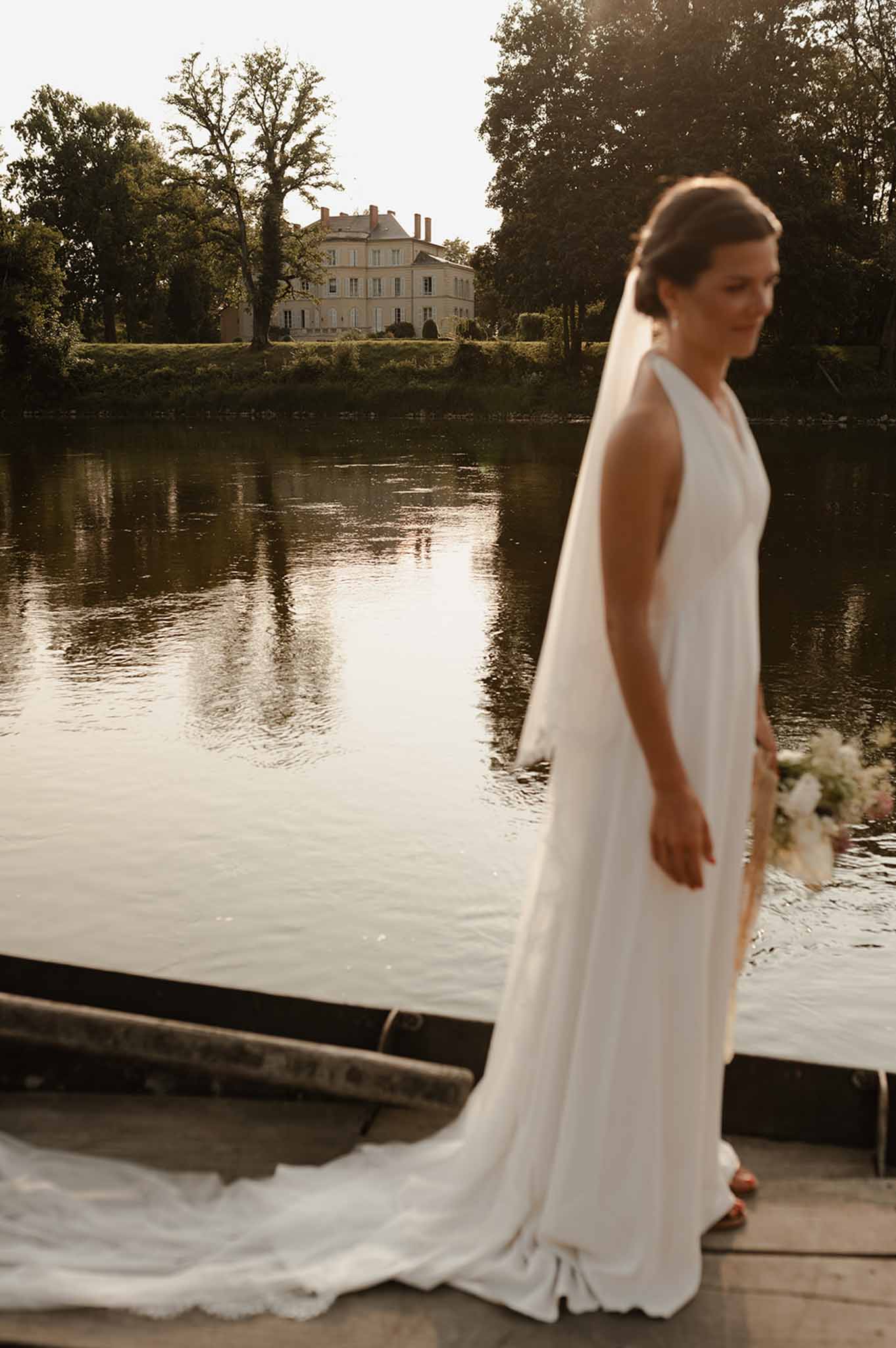 Bride in ivory column gown and long veil standing on dock, classical château reflected across river in background