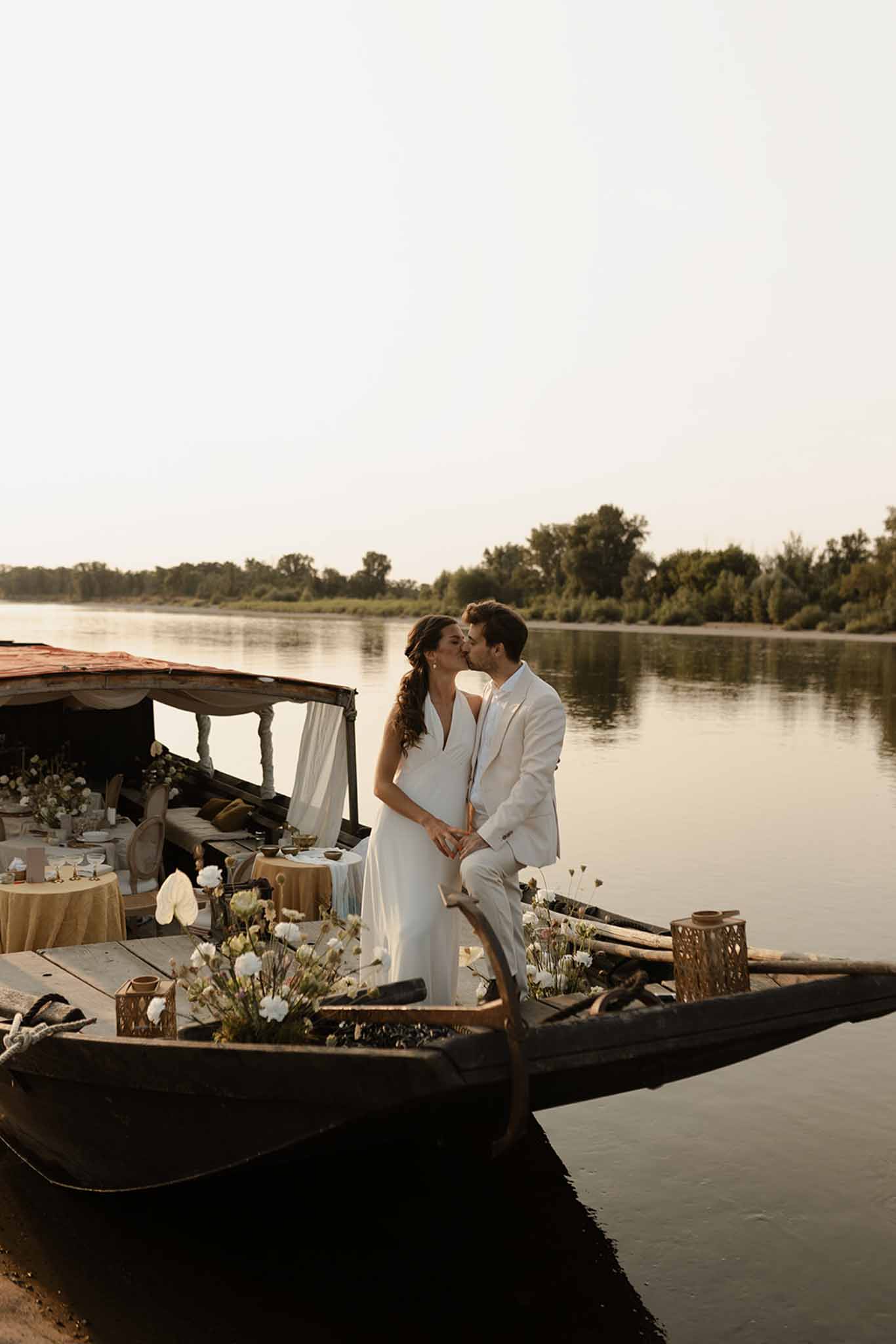 Bride and groom on decorated wooden boat on river, dried white flowers in dark vessels, canvas canopy reception setup behind