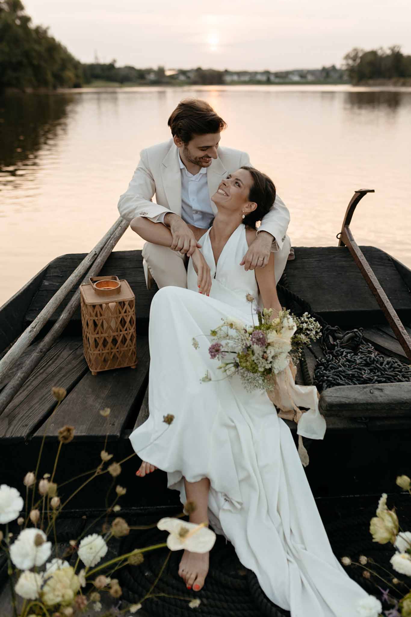 Bride and groom seated in wooden rowboat on calm river with white and mauve bouquet and rattan basket