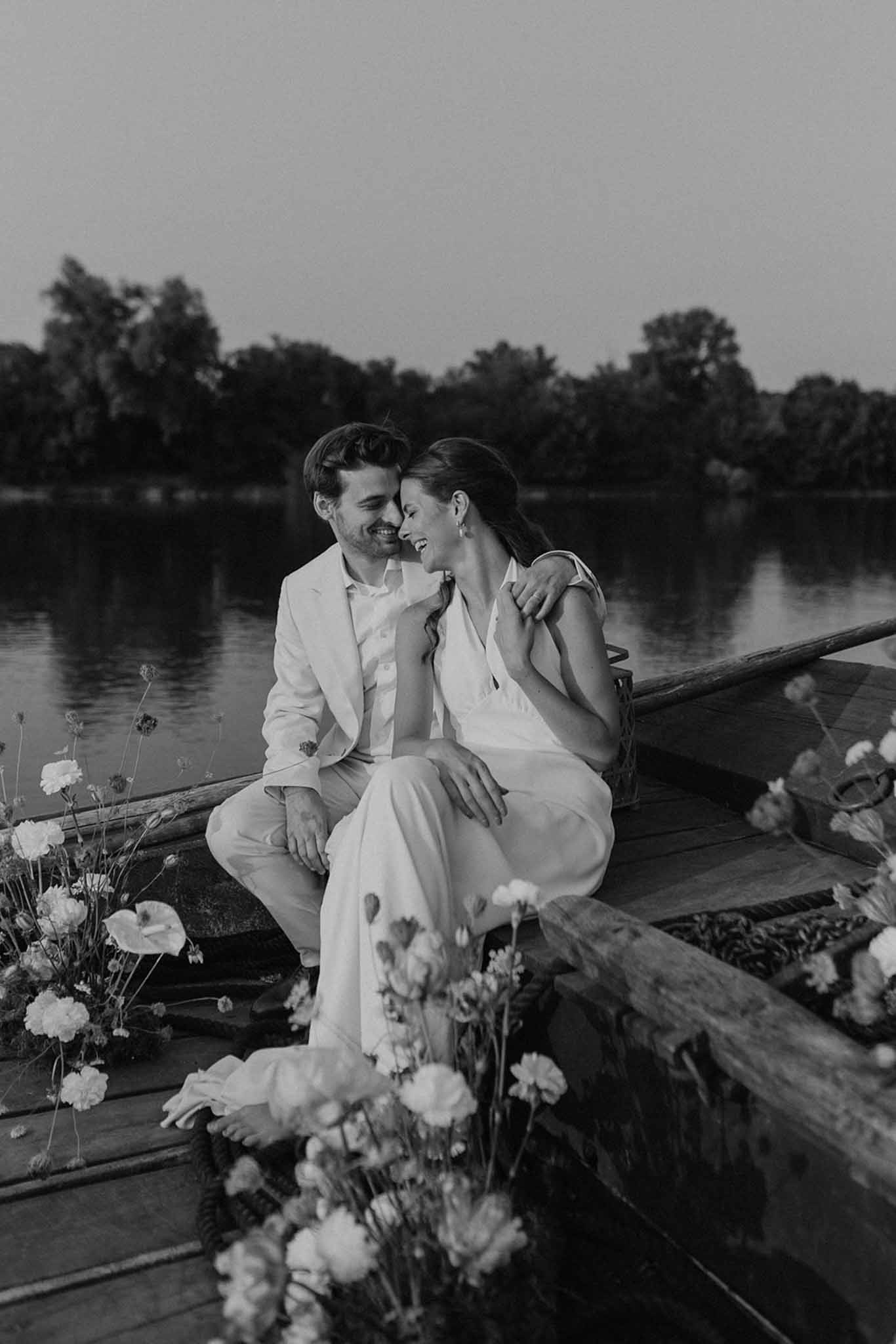 Black and white portrait of bride and groom seated by a river with baby's breath floral arrangements