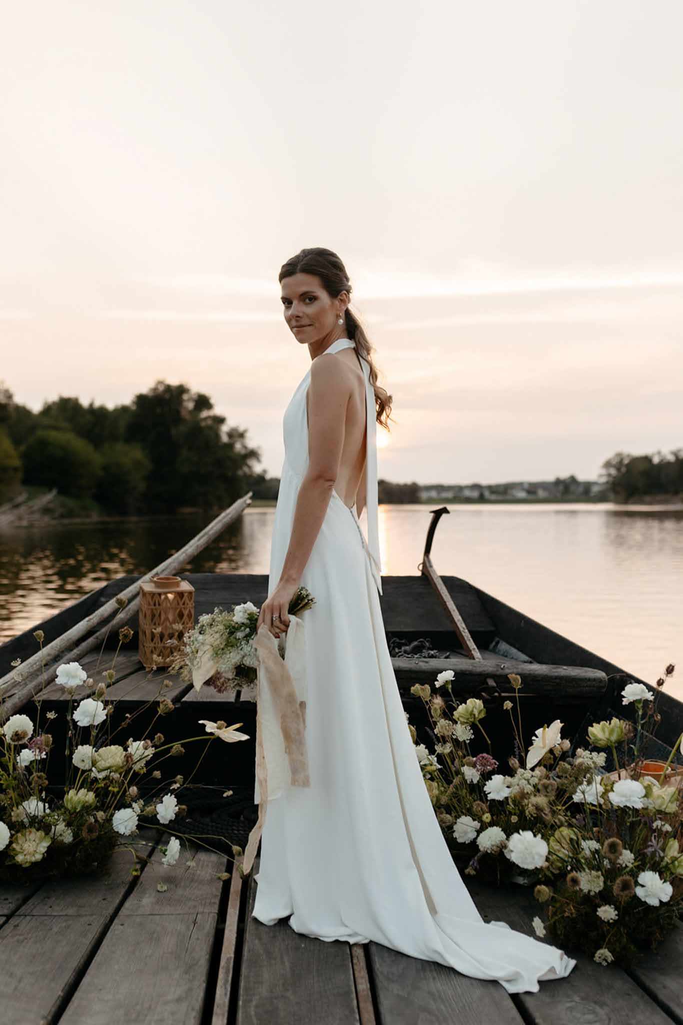 Bride posing on wooden boat at dusk wearing white open-back dress with carnation and dried floral decorations