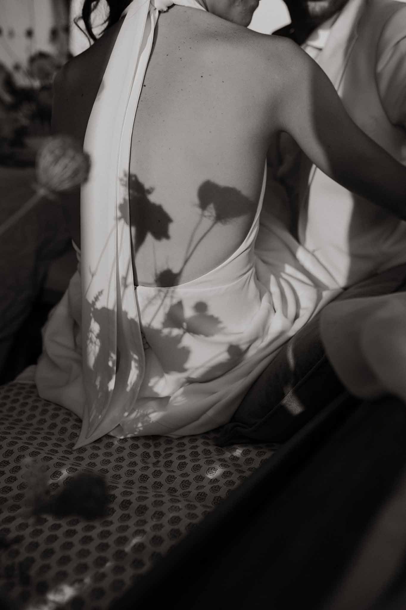 Black and white close-up of the open back of a bride's dress showing sheer panels casting floral shadows during preparation.