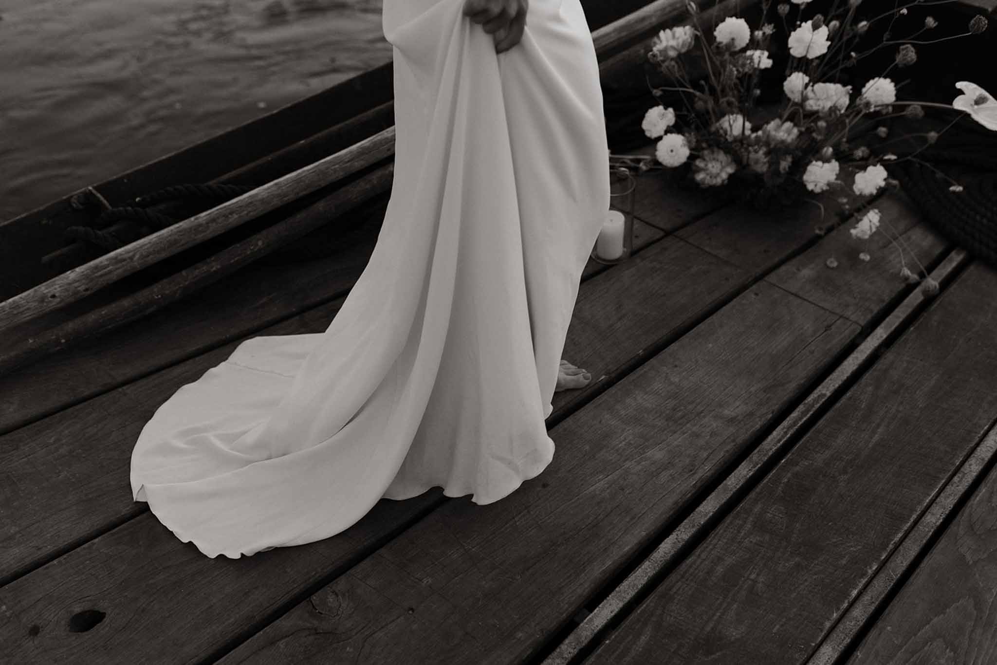 Black and white detail of bride's flowing train draped across weathered wooden deck beside white flowers