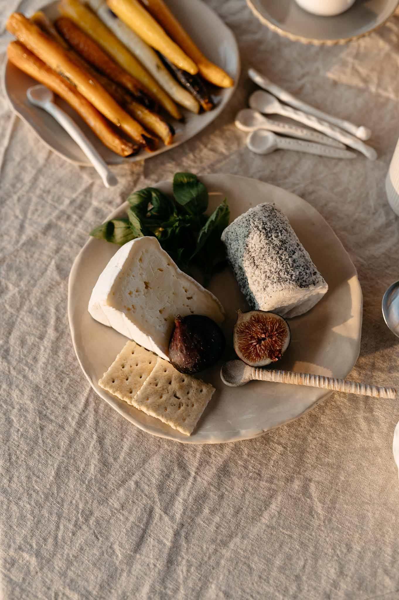 Overhead detail of cheese board with figs, roasted carrots, and white ceramic plates on linen