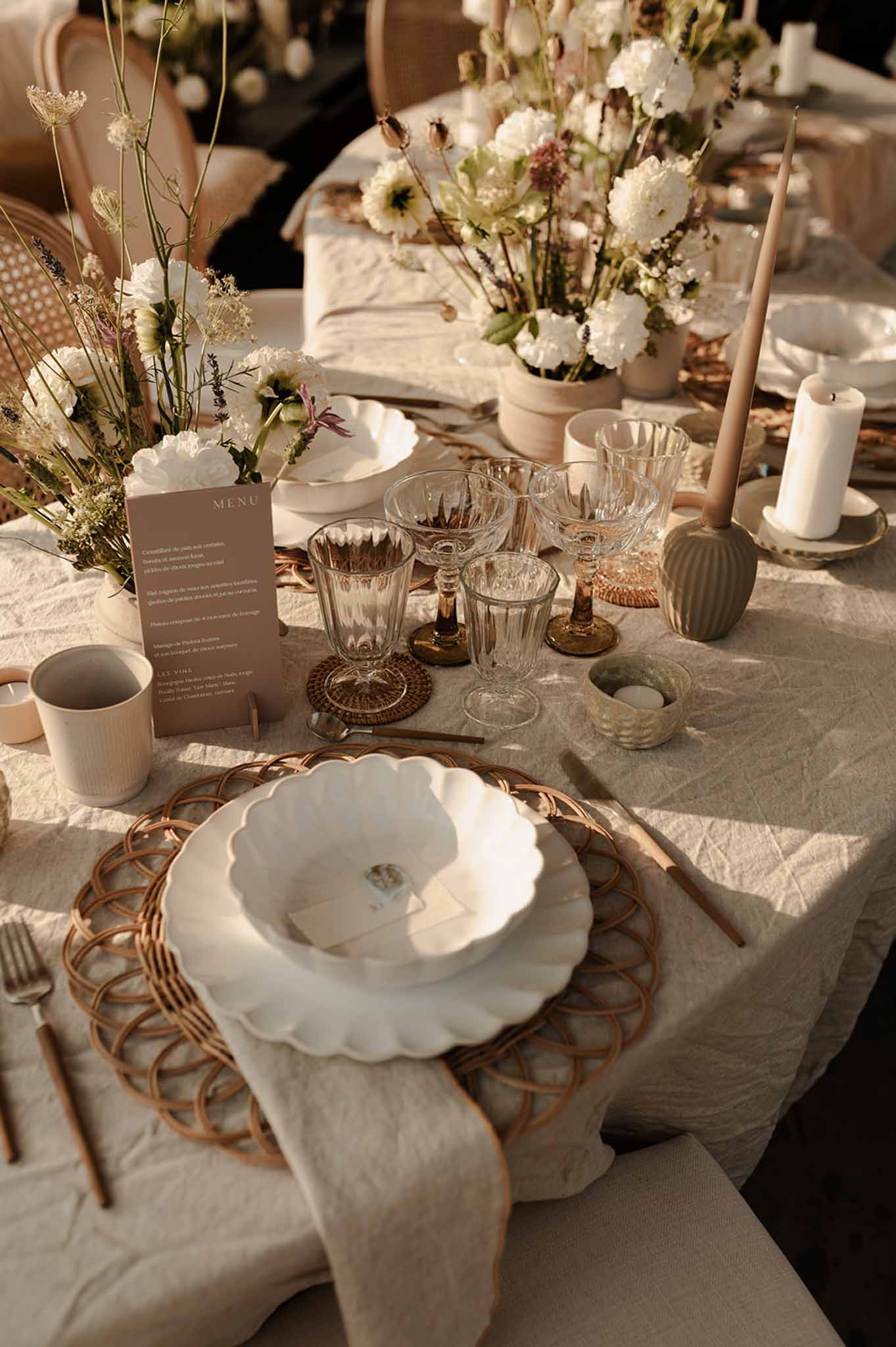 Close-up place setting with scalloped white plate on rattan charger, cream napkin, and white daisy centerpieces