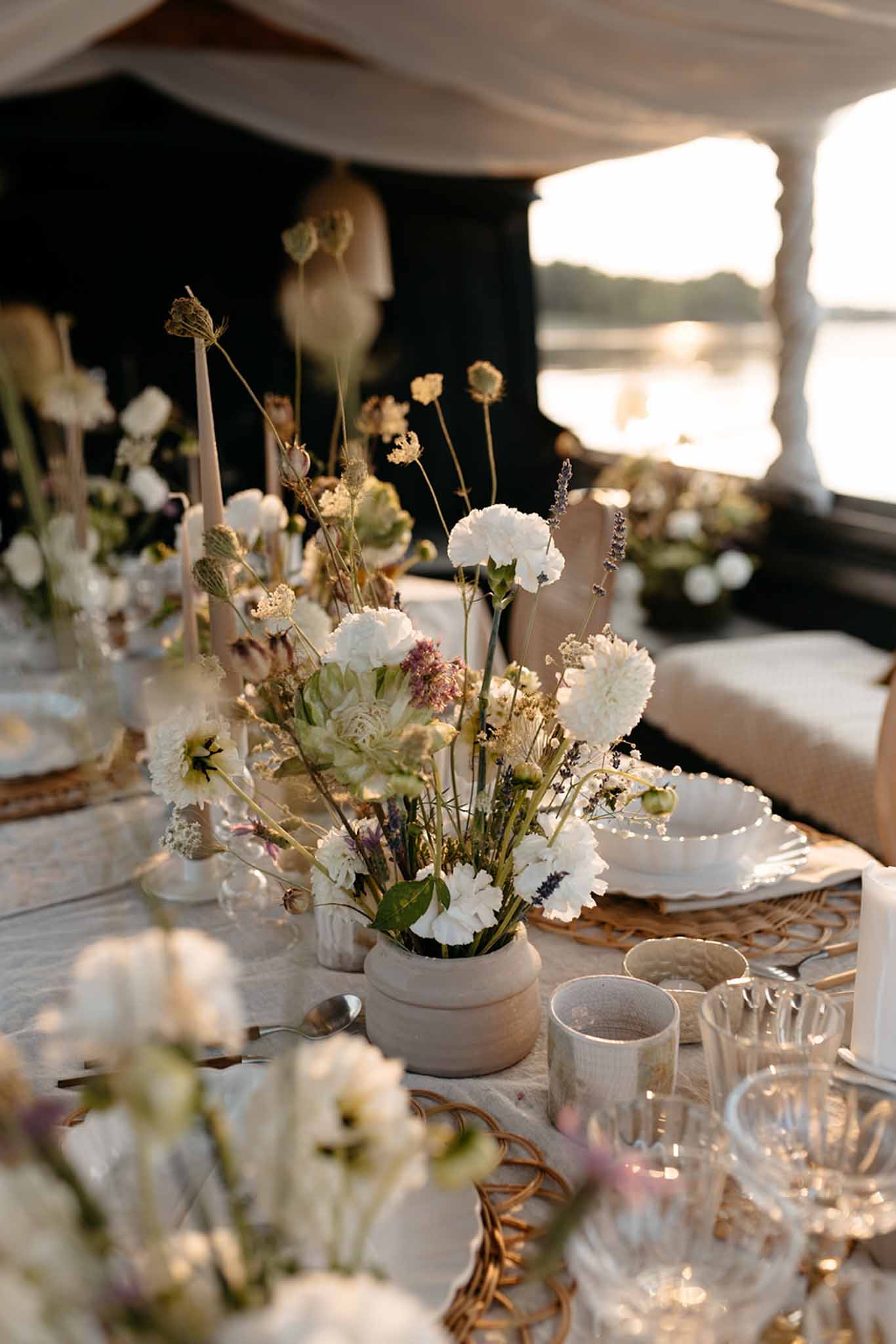 Reception table with white ceramic vessels of carnations and dried flowers under a marquee with water views