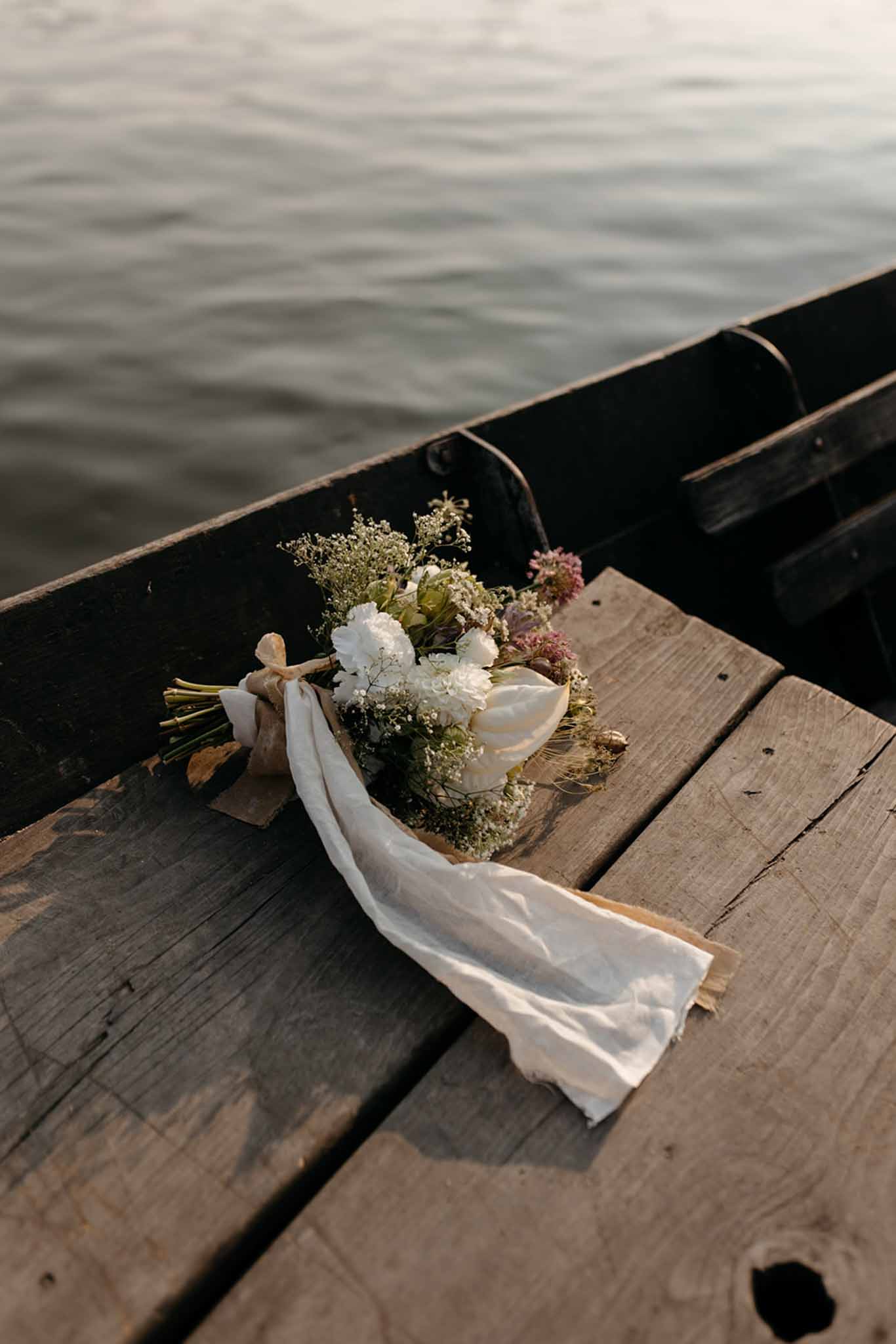 Bridal bouquet of white peonies and dusty mauve dried flowers resting on weathered dock by boat