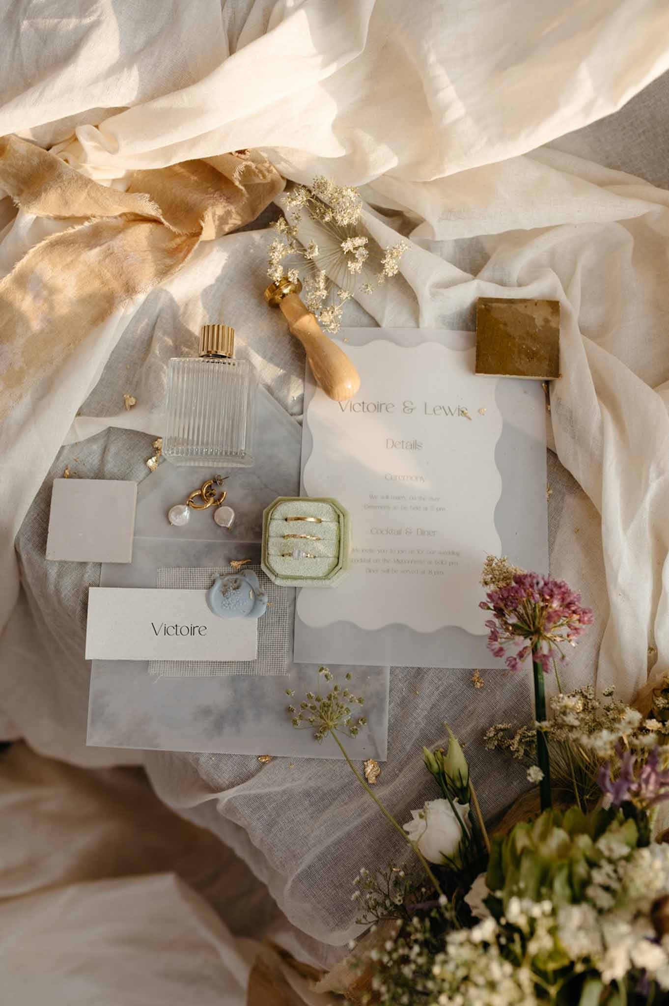 Flat lay of wedding stationery, perfume, ring boxes, gold accessories and a hand-tied bouquet of cream and burgundy florals.