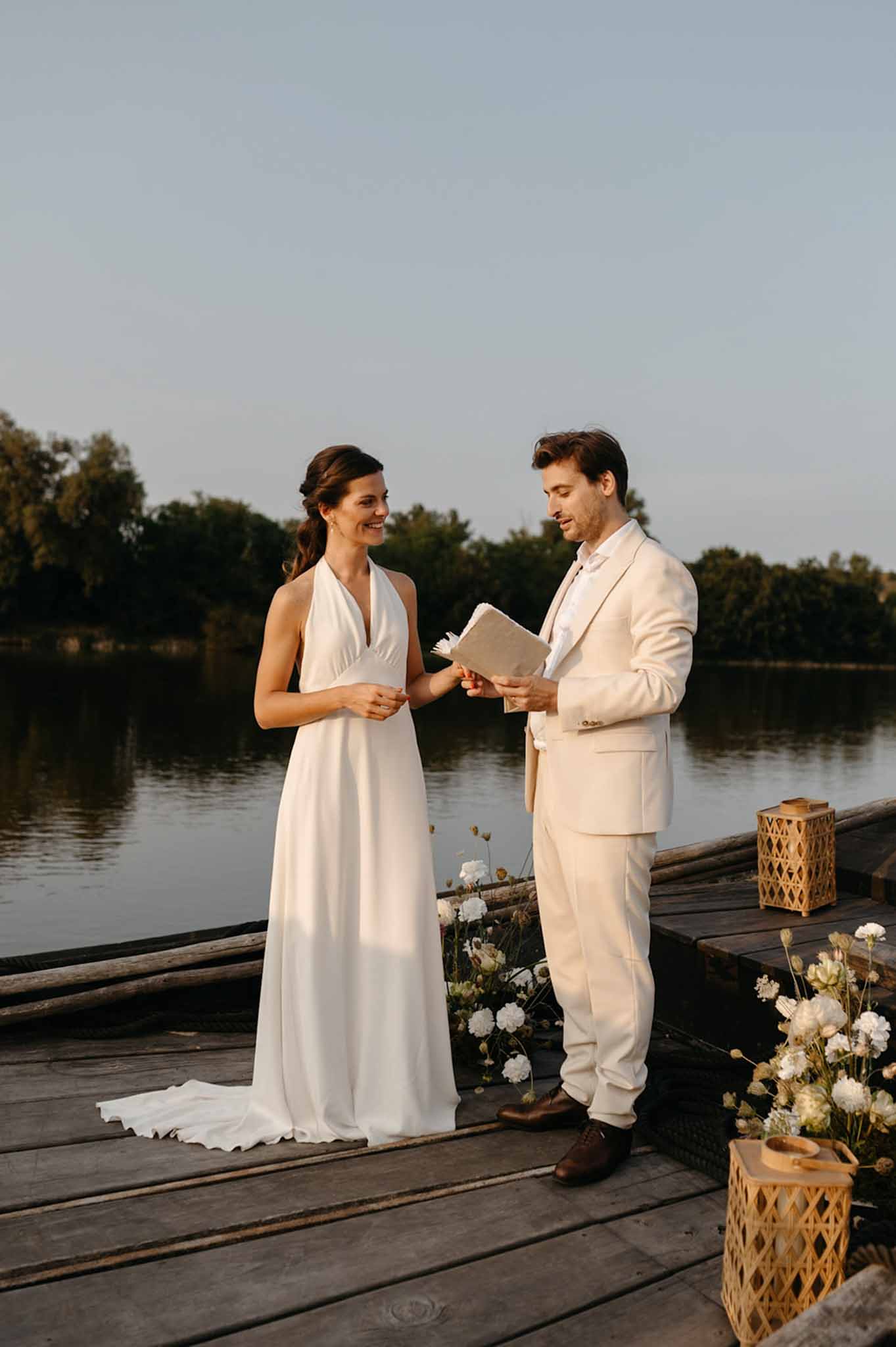 Bride and groom exchanging vows on wooden riverside dock, groom holds vow notebook, dried white flower baskets flanking