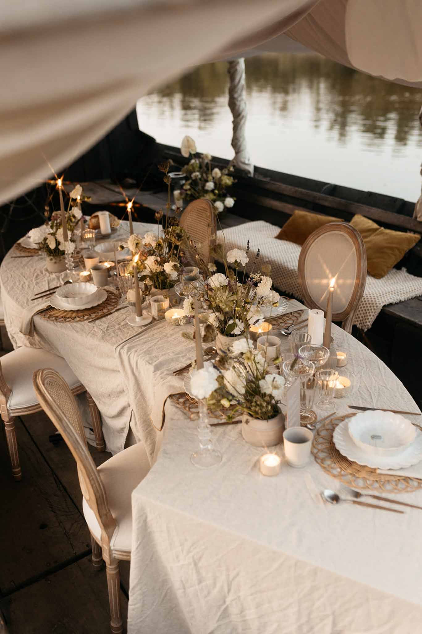 Reception table in marquee with cream linens, gold chargers, white dahlias, dried grasses, and rattan chairs by water