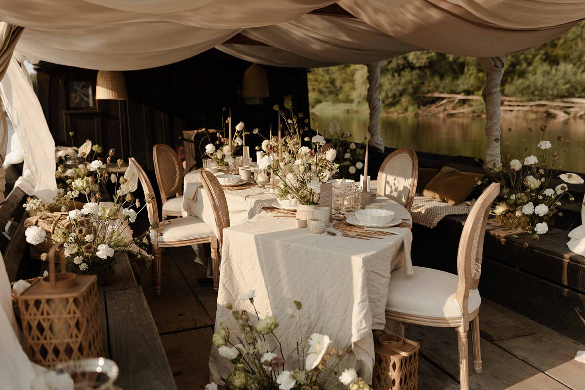 Outdoor reception under canvas tent on wooden deck, round tables with cream linens, rattan chairs, white carnation vases