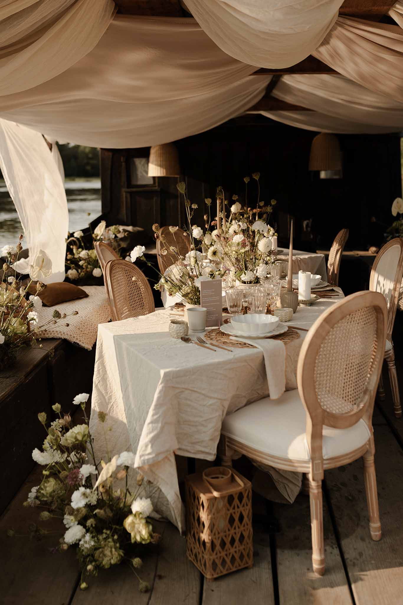 Long banquet table under ivory draped tent with jute runner, white flower centerpieces and rattan chairs
