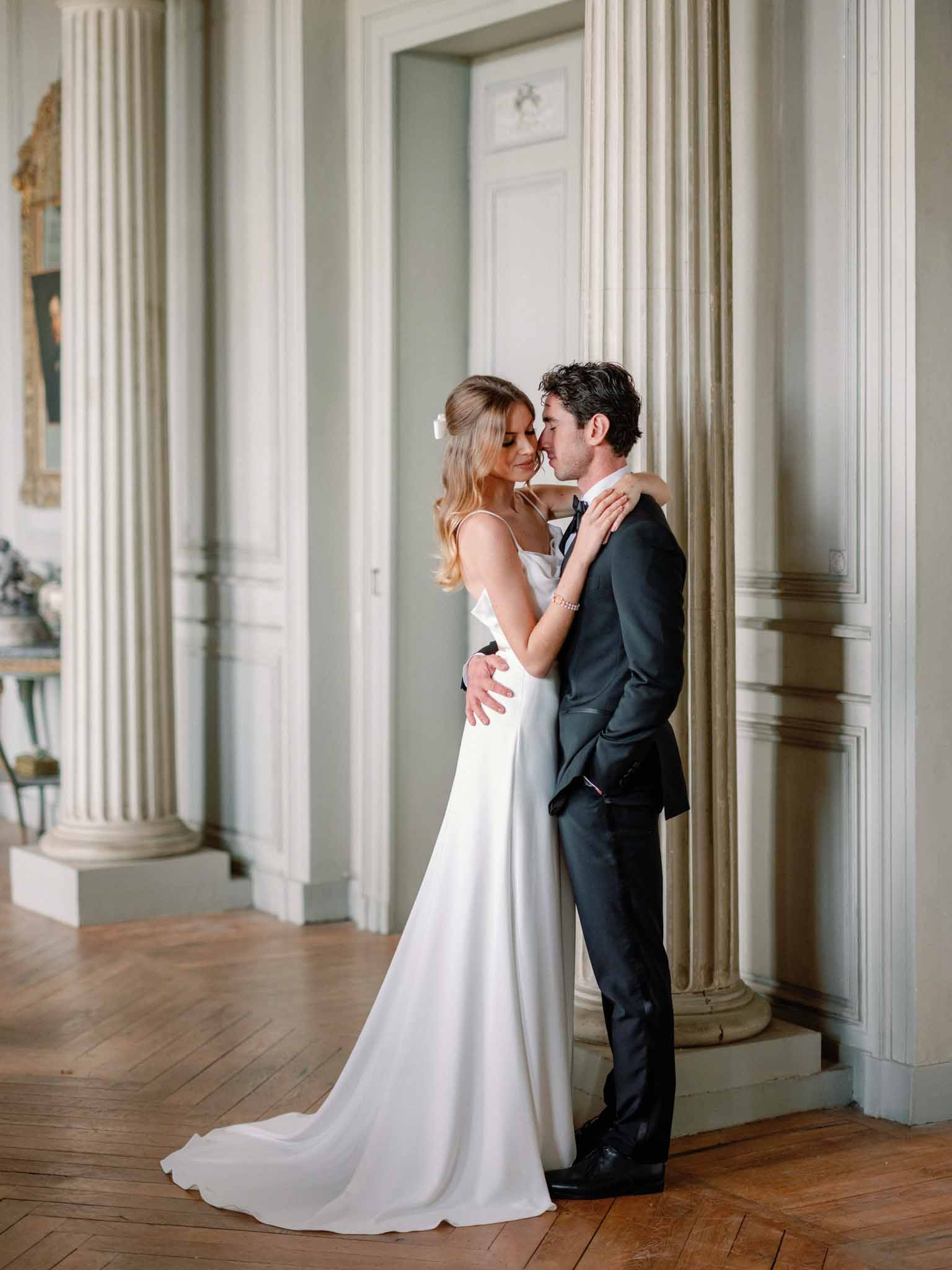 Bride and groom embracing in classical hallway with tall white fluted columns, pale green walls and herringbone wood flooring