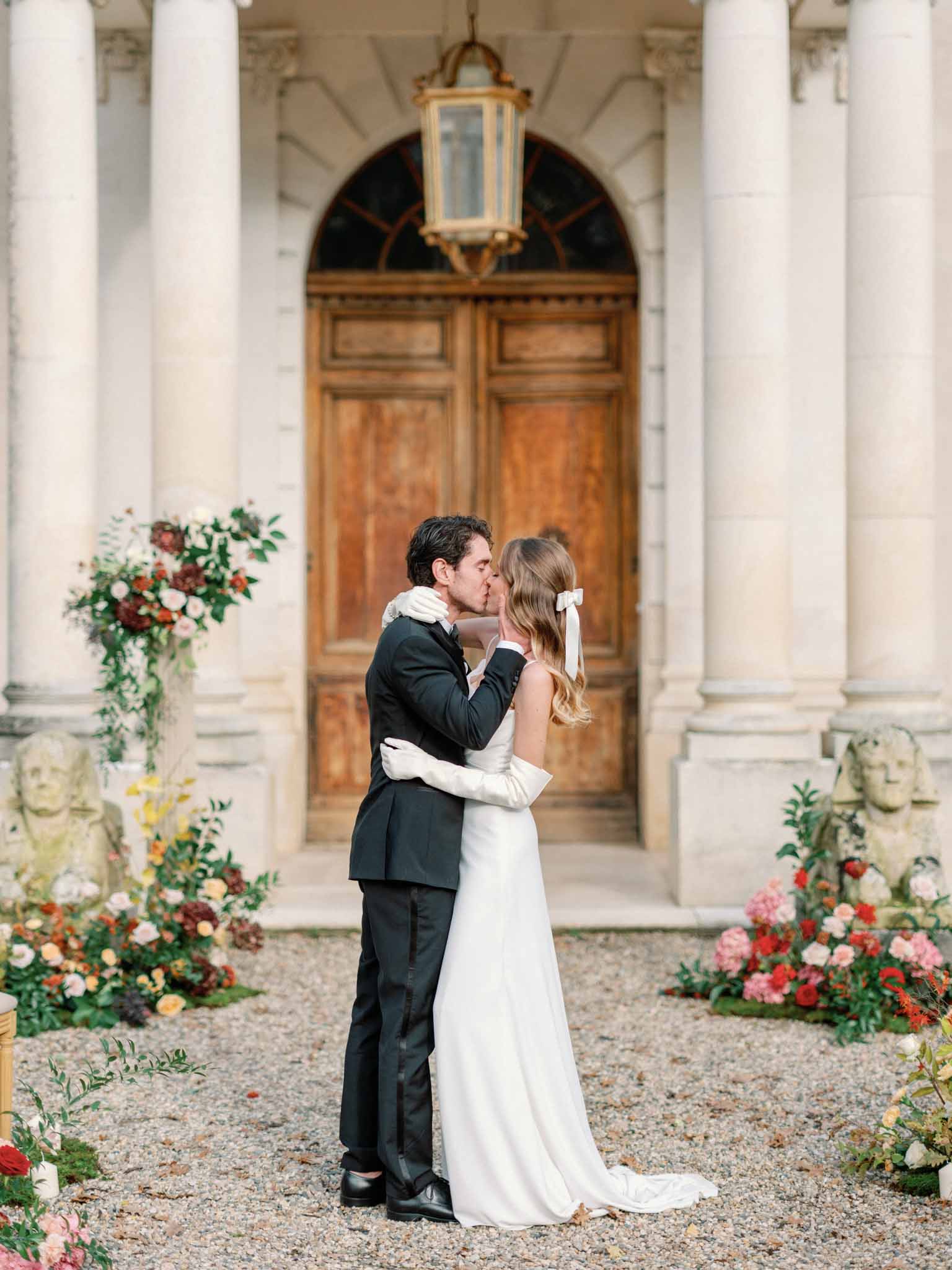 Bride and groom kissing under columned portico with coral and blush floral arrangements at classical courtyard venue