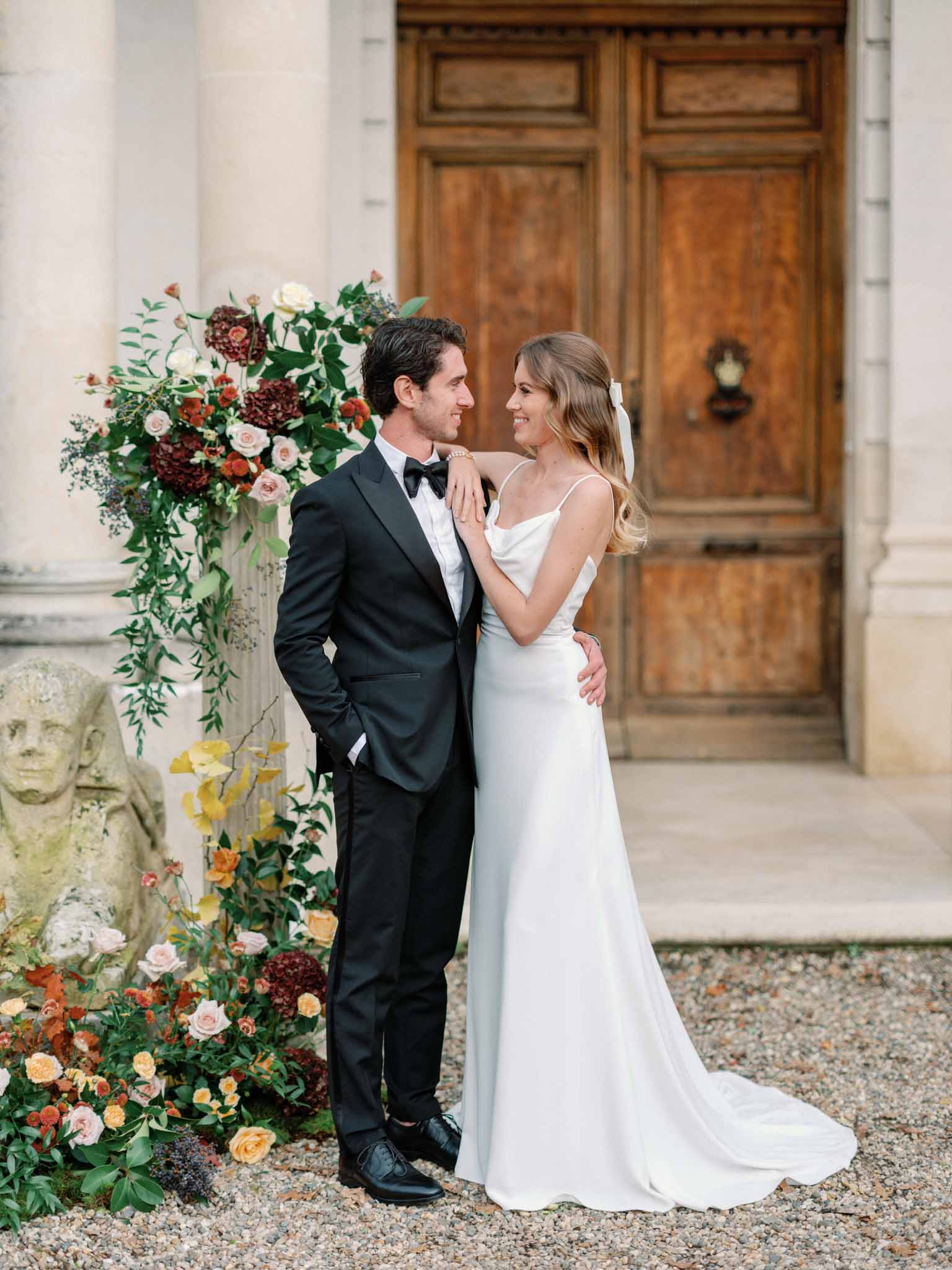 Bride and groom beside ornate door framed by burgundy dahlia and blush rose floral installations in courtyard