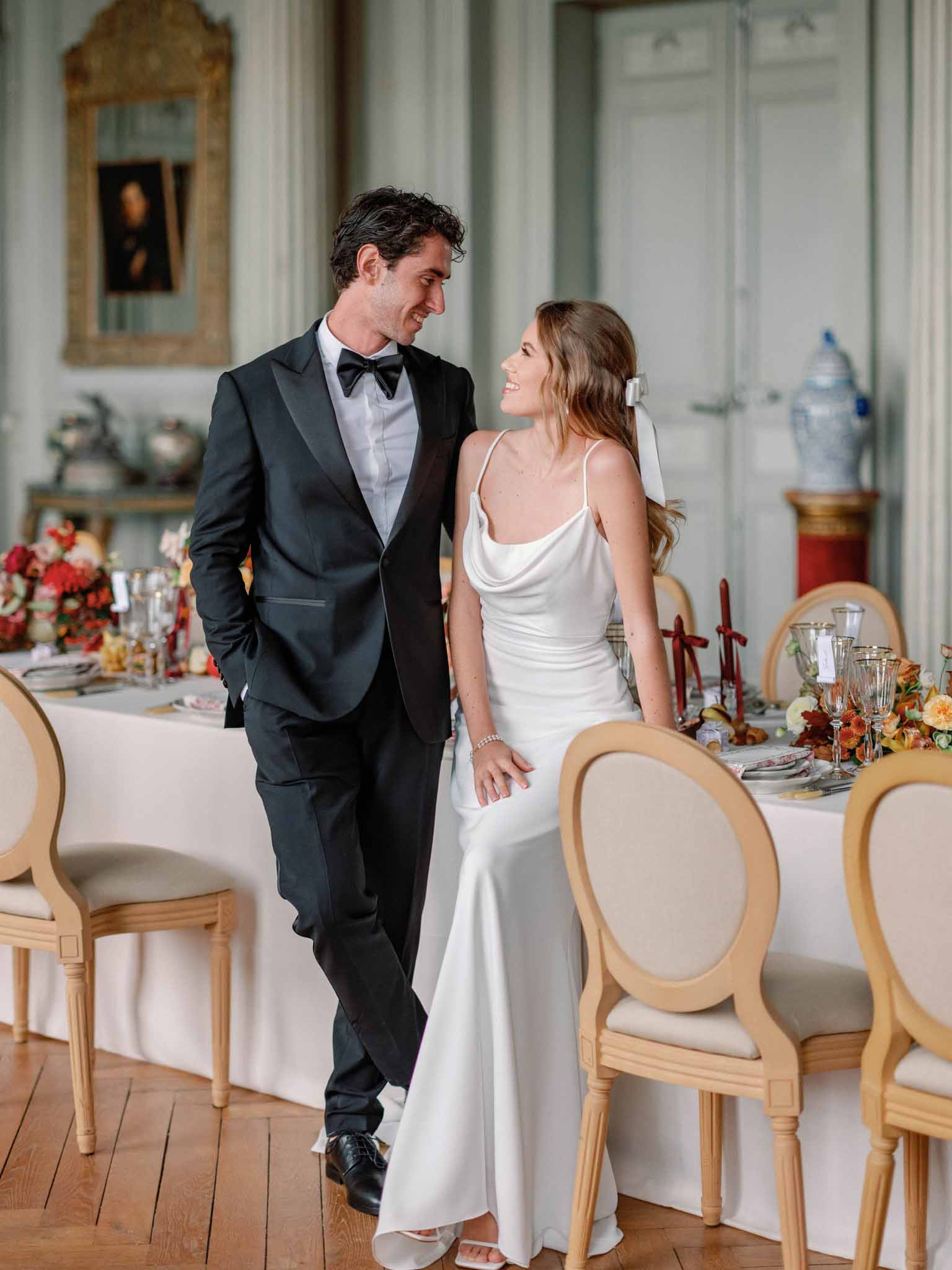 Bride in ivory slip dress and groom in black tuxedo walk through neoclassical reception room with autumn floral centerpieces