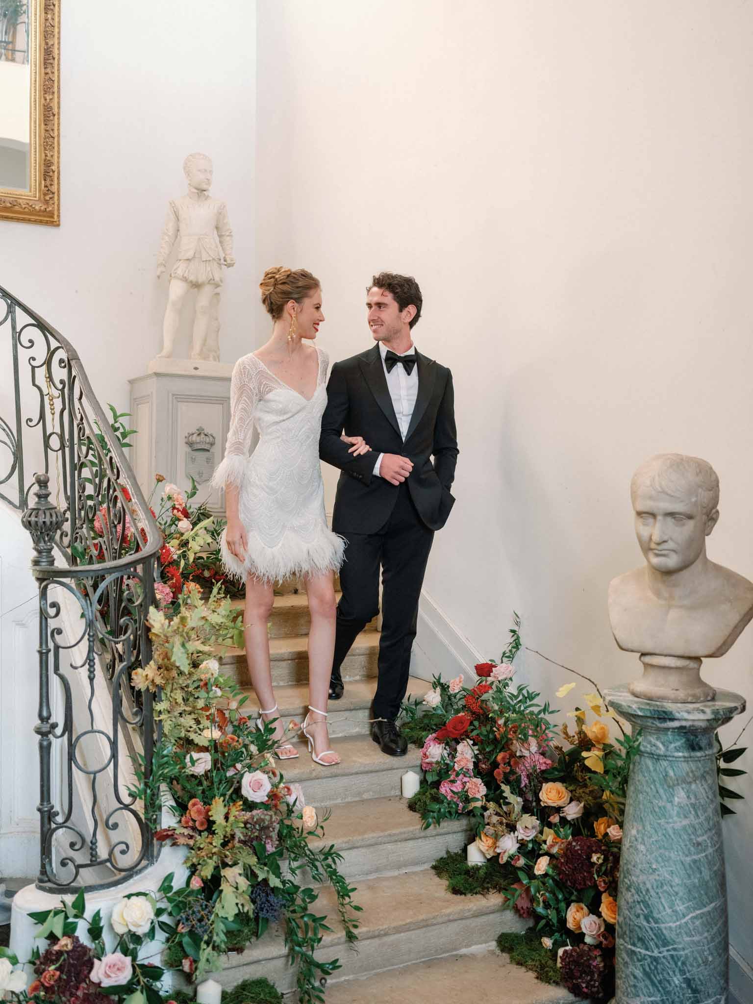 Bride and groom descending interior staircase lined with coral and burgundy floral arrangements in classical gallery