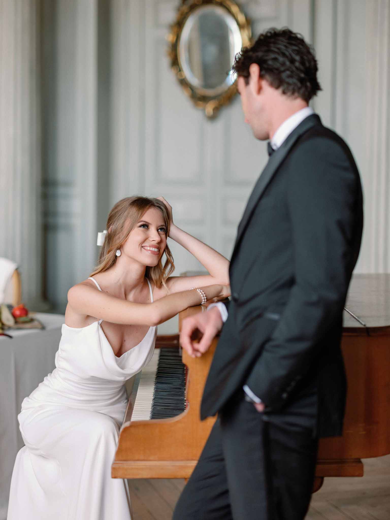 Bride at piano in white draped gown, groom in charcoal suit beside her, gold-framed mirror on grey paneled wall behind