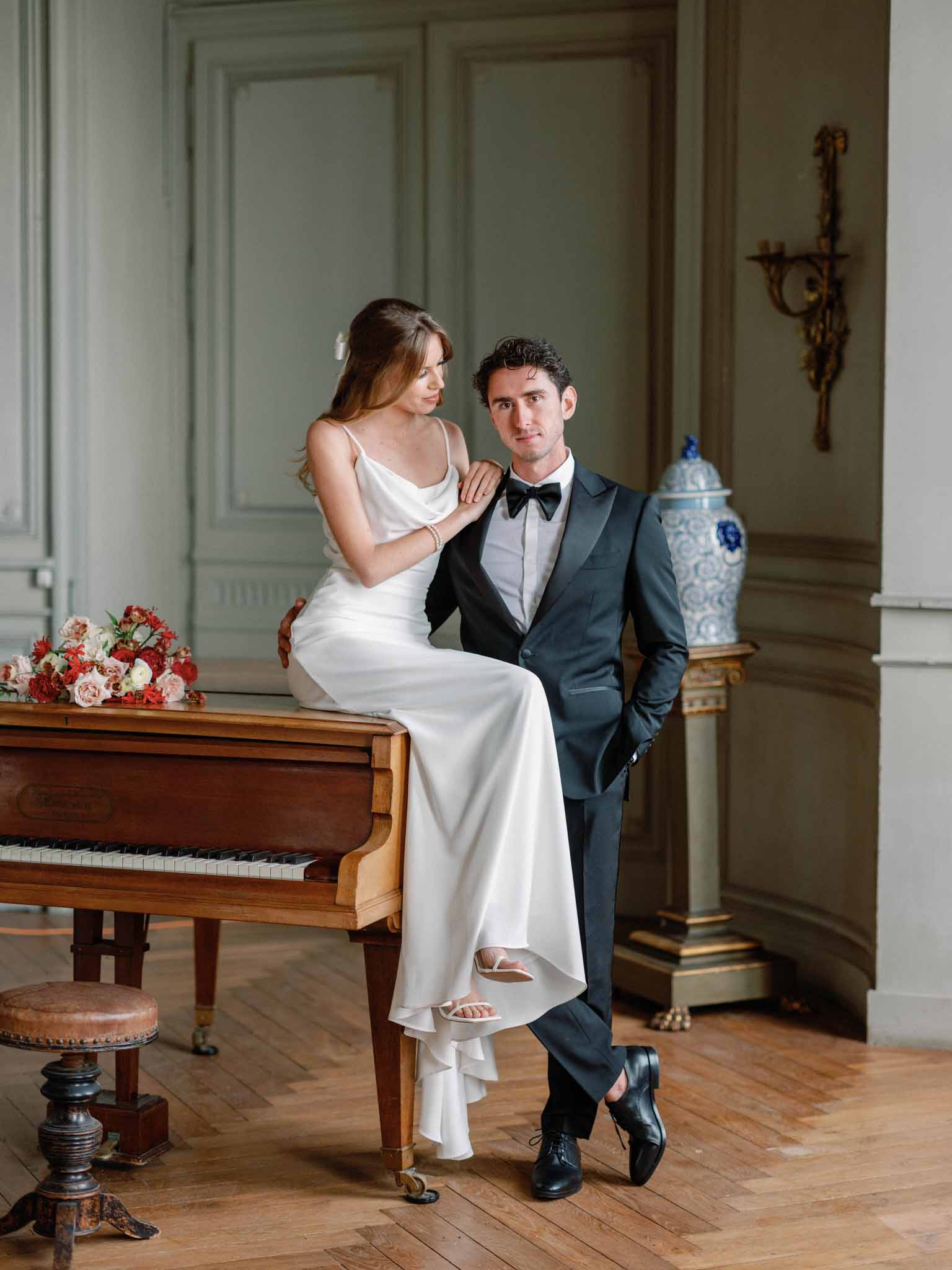 Bride seated on piano with groom beside her in sage green paneled room with red rose bouquet