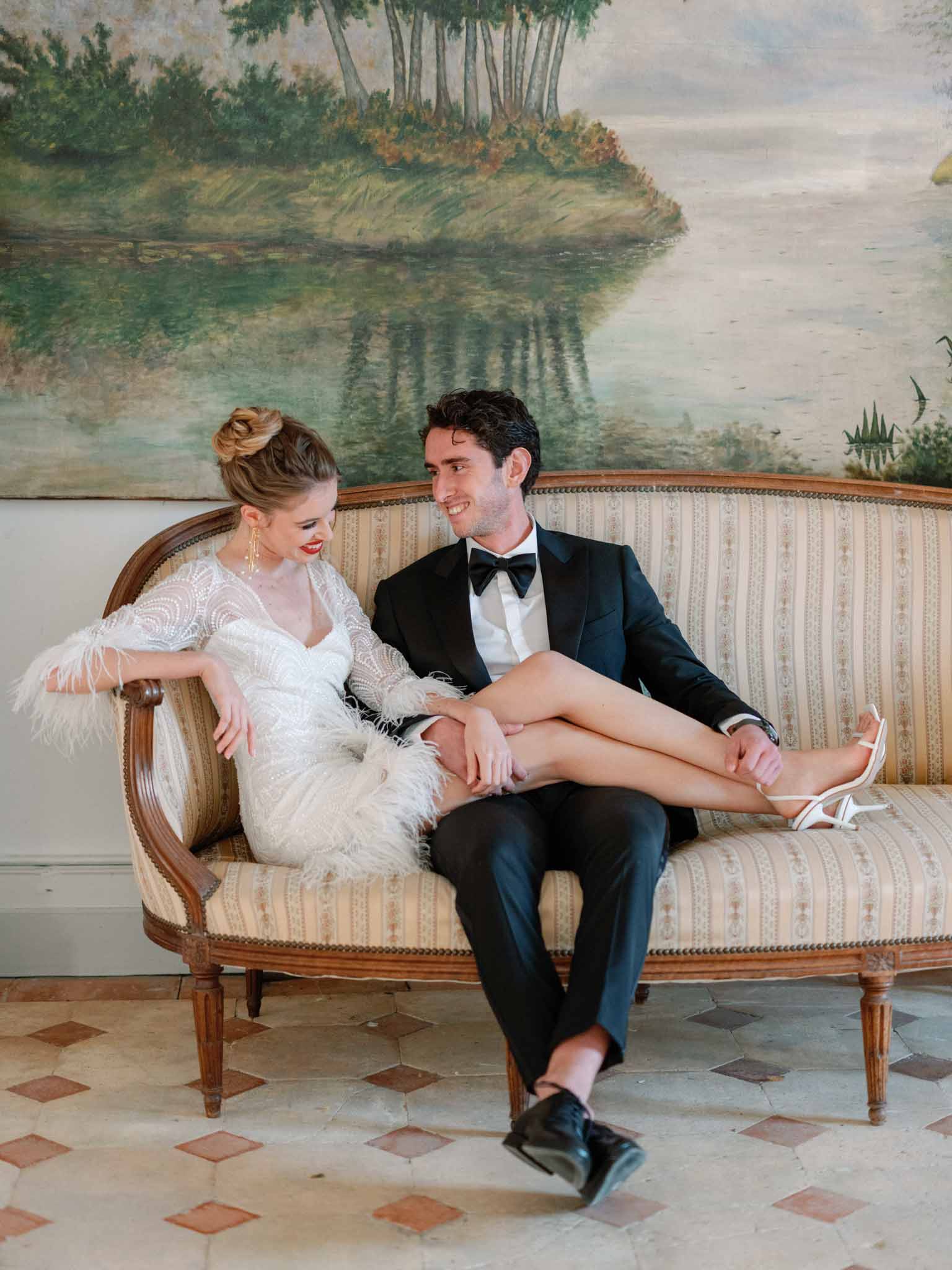 Bride in beaded sheath dress and groom in black tuxedo posing on period sofa in front of landscape painting