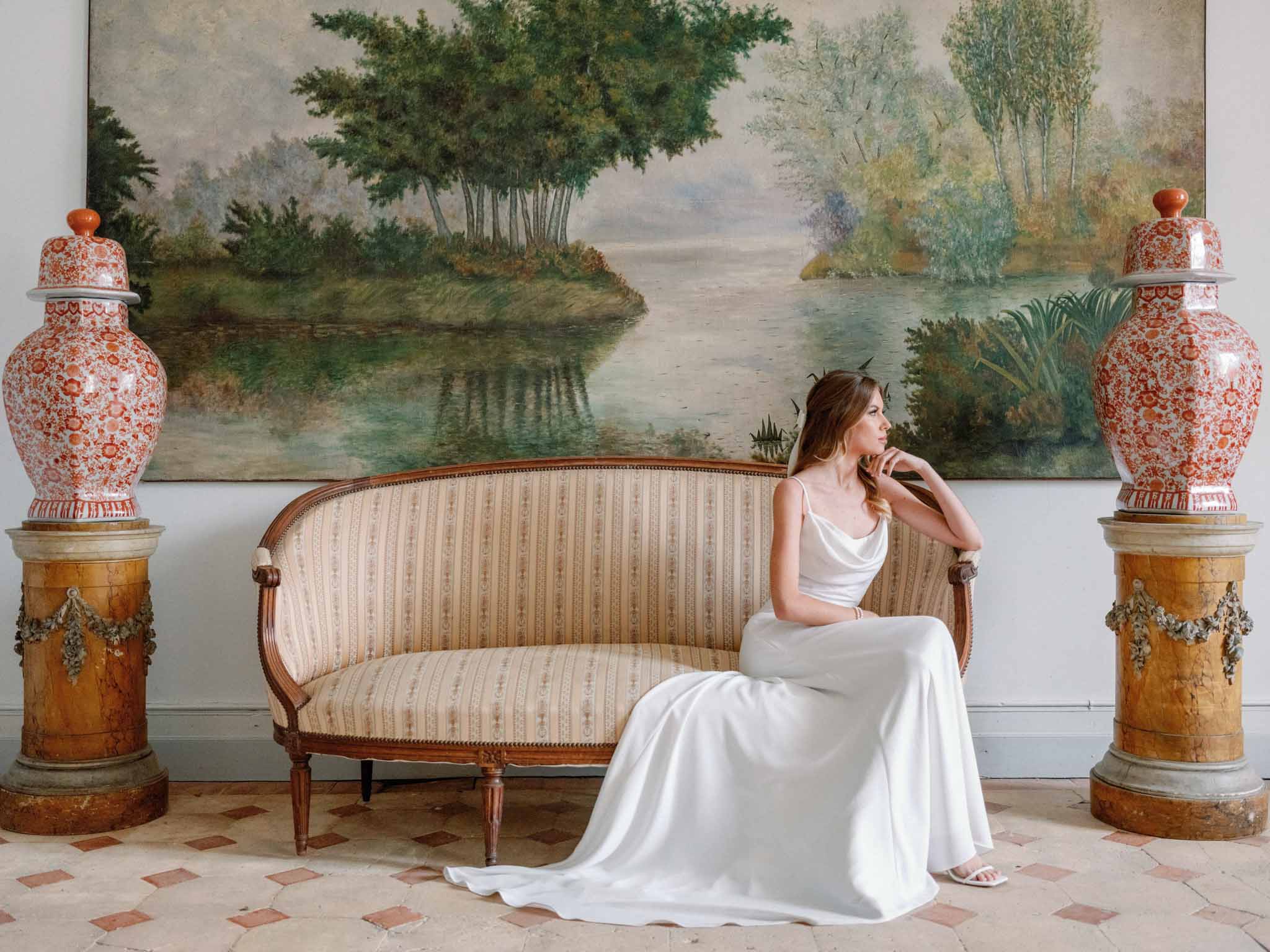 Bride in white cowl-neck dress seated on striped settee with chinoiserie ginger jars