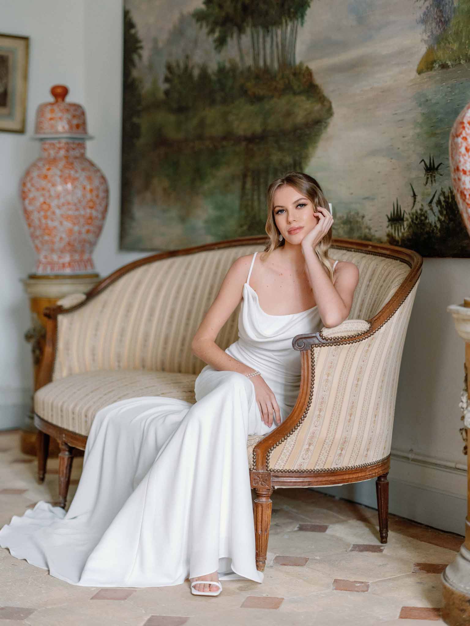 Bride seated on cream striped armchair in neoclassical room wearing white cowl-neck dress with coral ceramic decor